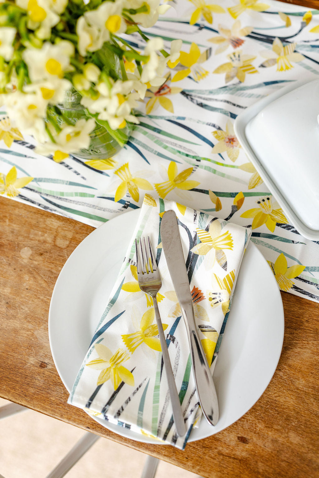 Table setting with Daffodil floral-patterned napkins, fork, knife, and white plate on a wooden table.