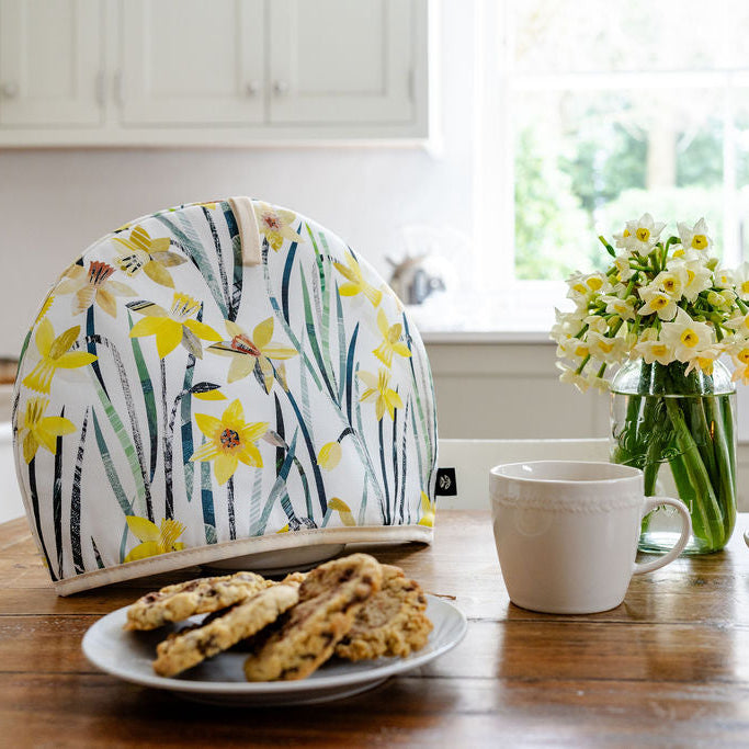 A Daffodil Tea Cosy has been placed on a wooden table in a cream kitchen with  plate of cookies a cup of tea and small vase of daffodils.