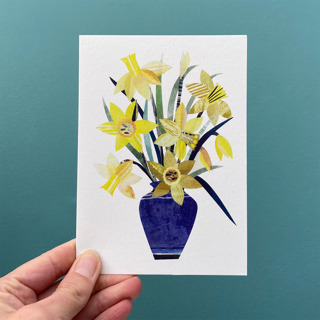 A greeting card featuring a blue vase of yellow daffodils is being held in front of a blue green wall.