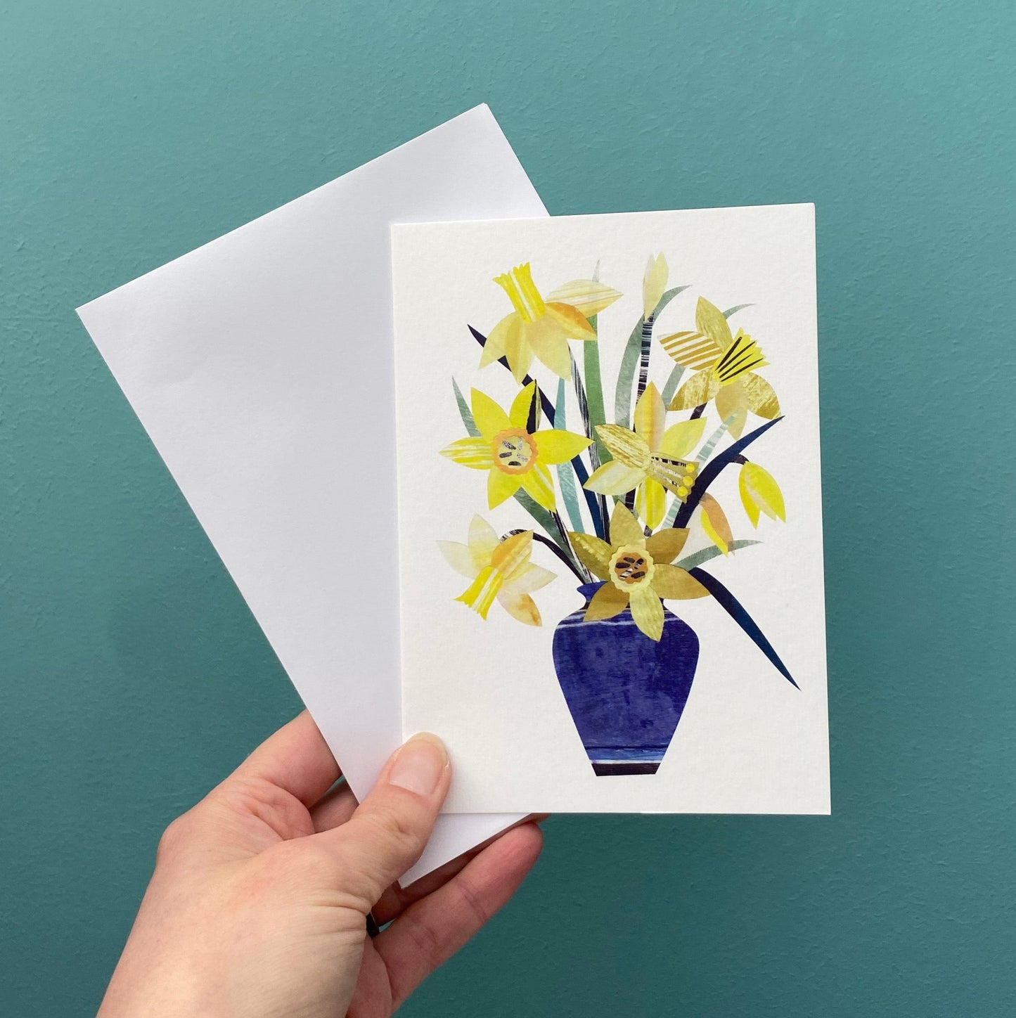 A greeting card featuring a blue vase of yellow daffodils with a white envelope is being held in front of a blue green wall.