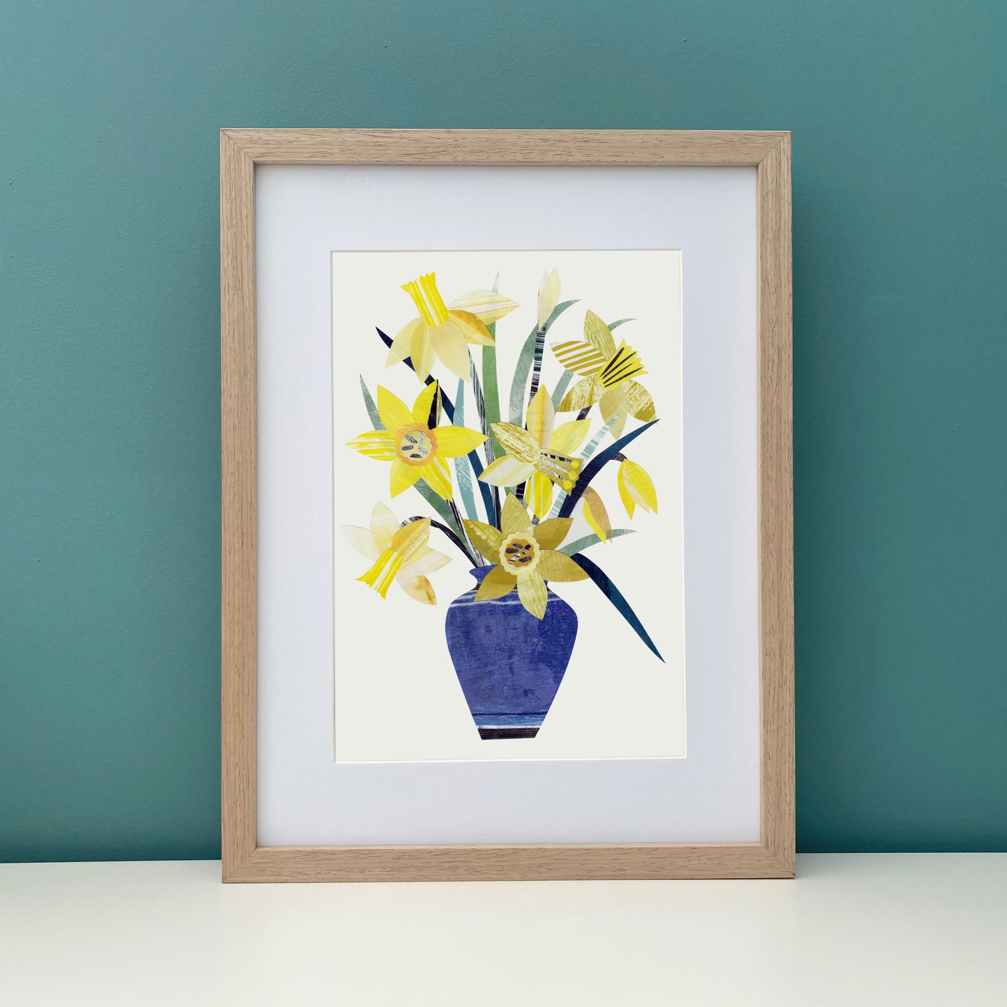 A Print of Yellow Daffodils in a Blue Vase has been placed in a wood frame against a teal wall.