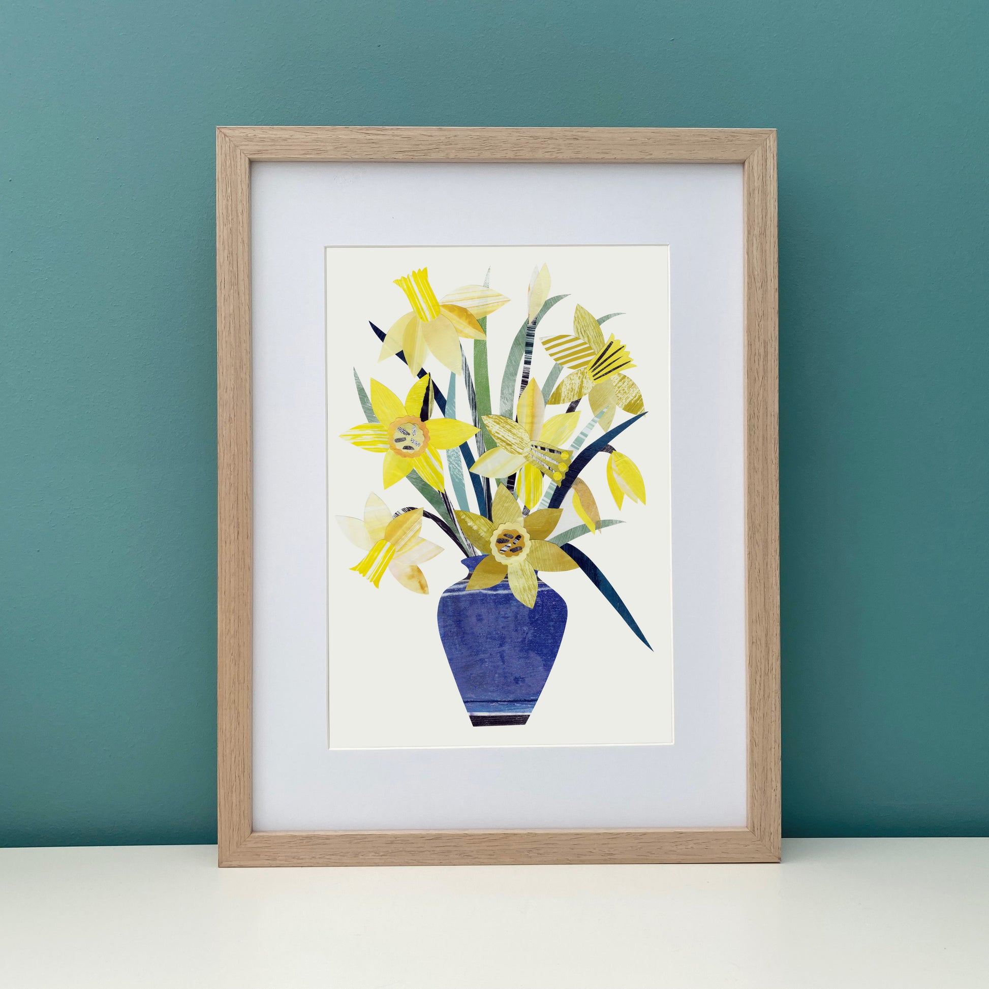 A Print of Yellow Daffodils in a Blue Vase has been placed in a wood frame against a teal wall.