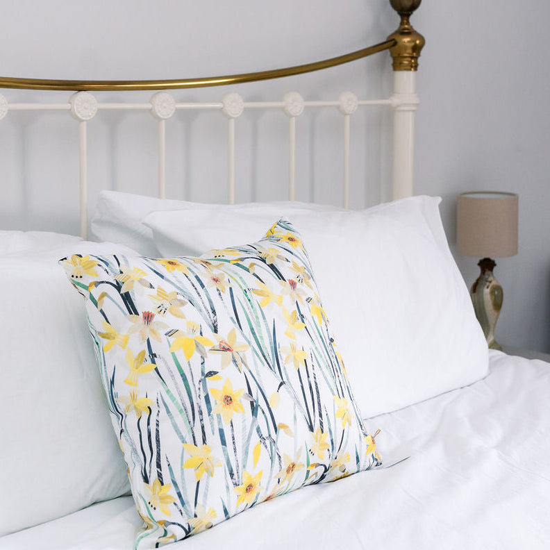 A white bed with a brass metal headboard, styled with white bedding and a decorative throw pillow featuring a yellow daffodil and green leaf pattern.