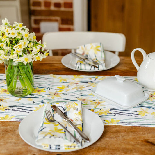 Dining table set with plates, cutlery, a vase of flowers, and a teapot with a Daffodil table runner and matching napkins.