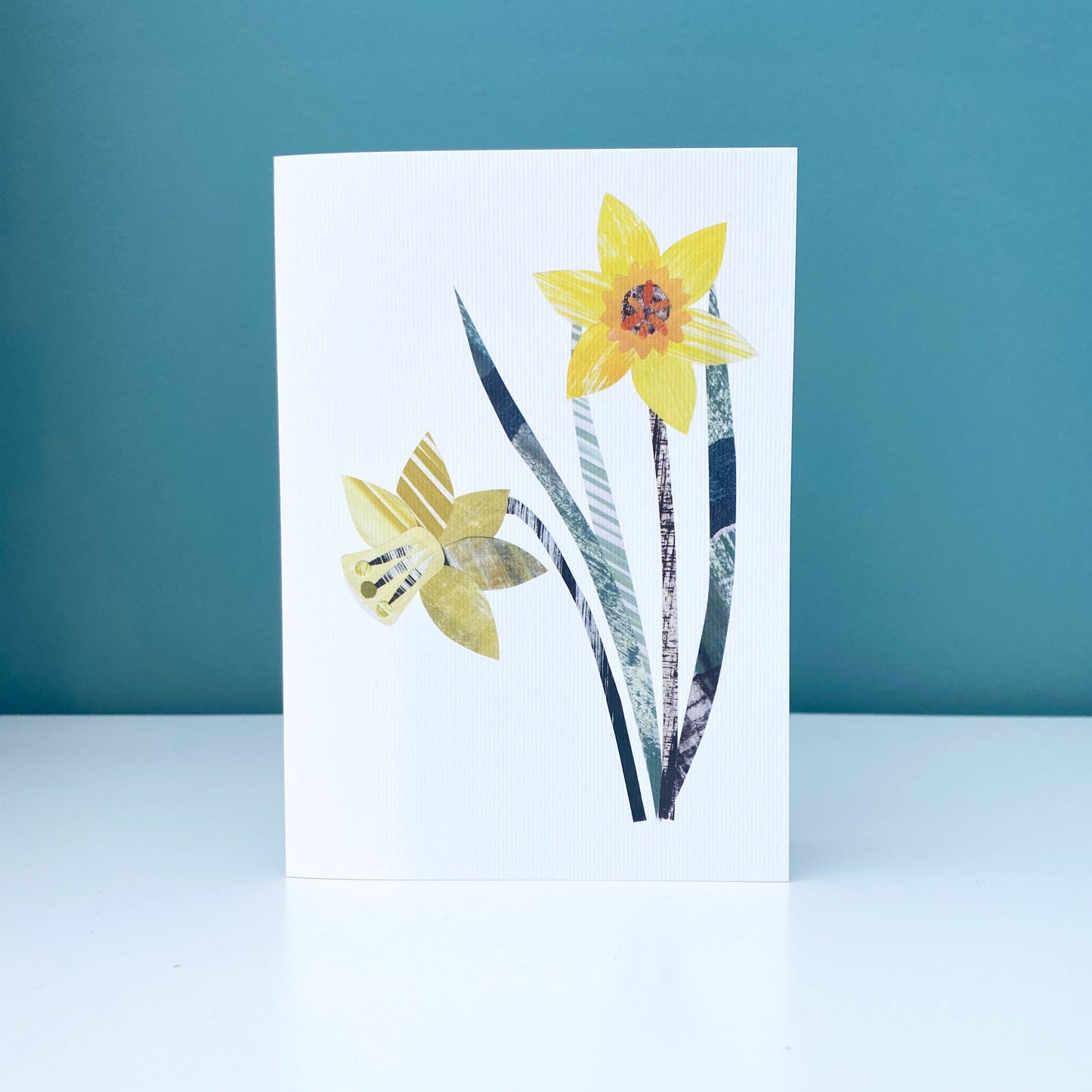 A Daffodil greeting card has been stood on a white shelf against a blue green wall.