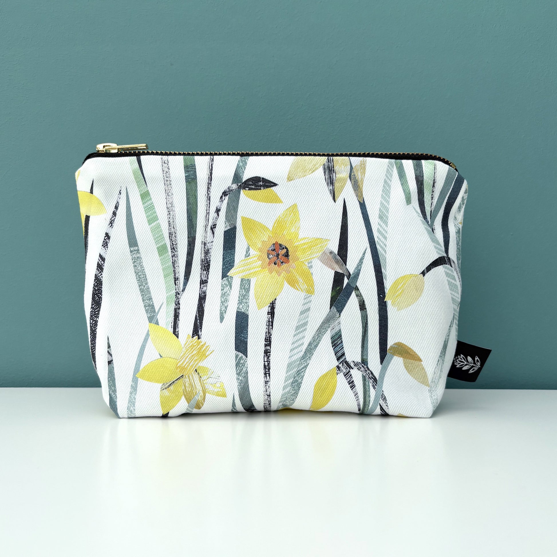 A Daffodil make up bag has been placed on a white shelf against a blue green backdrop.