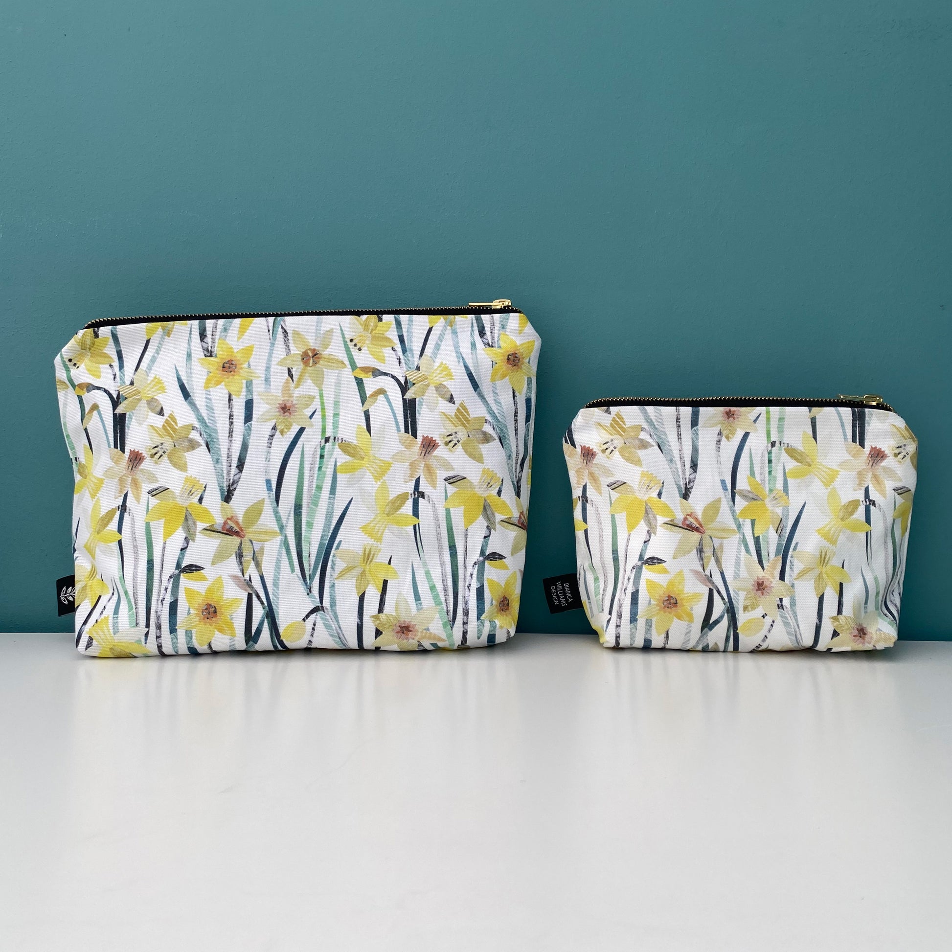 A large and small Daffodil wash bag have been placed side by side on a white shelf against a blue/green background.