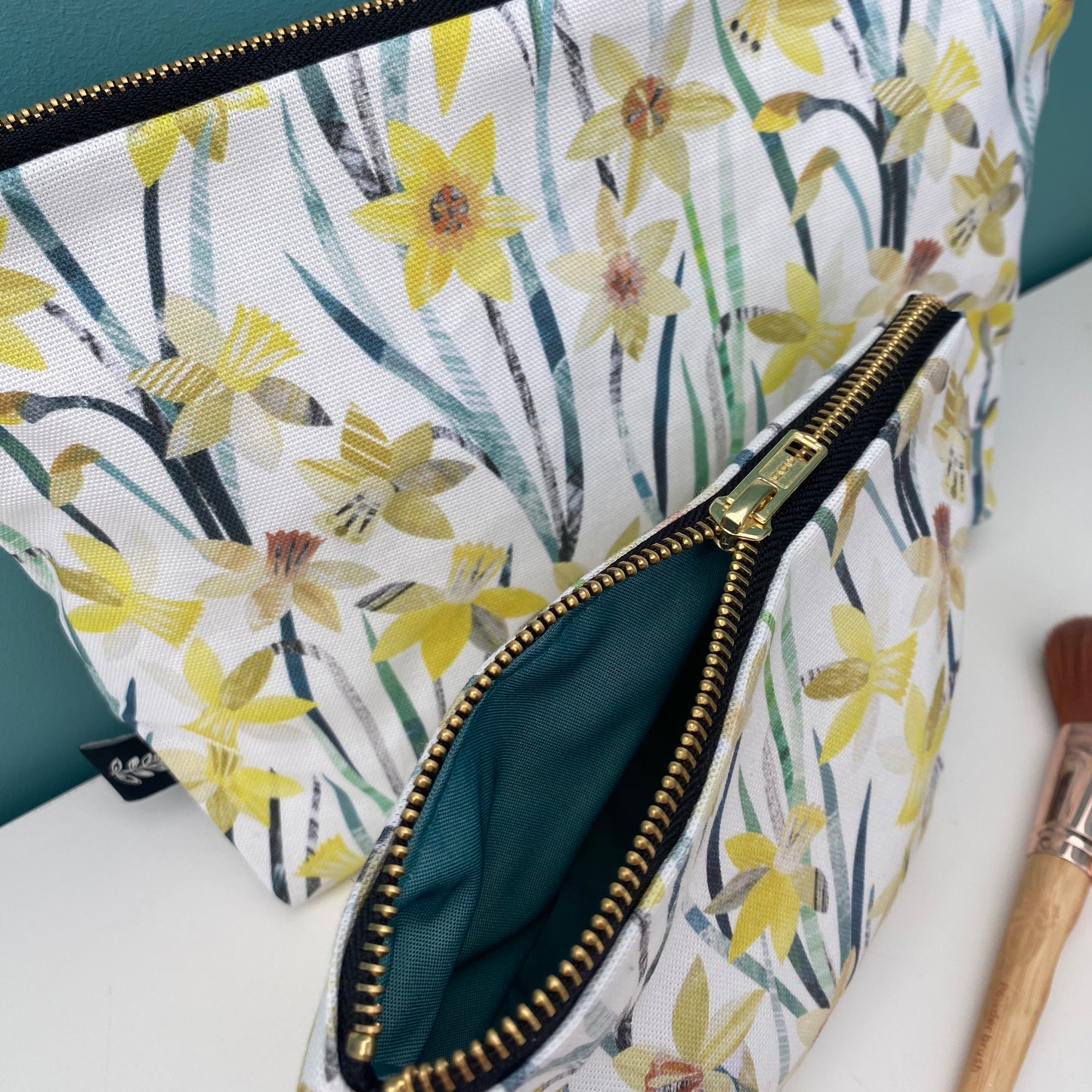 A small daffodil wash bag has been placed at an angle in front of a large daffodil wash bag.  The small wash bag has its gold zip half opened so that you can see the matching green lining inside.