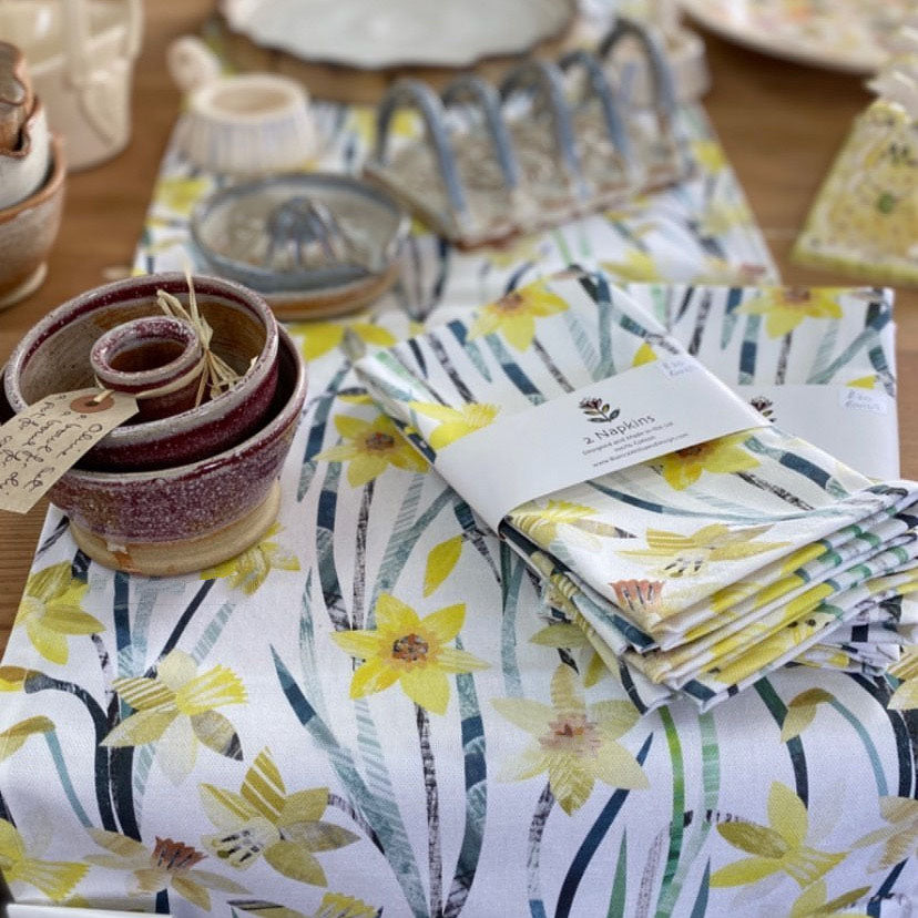 A Daffodil Table runner has been placed on a wooden table with some matching packaged napkins and some ceramic items.