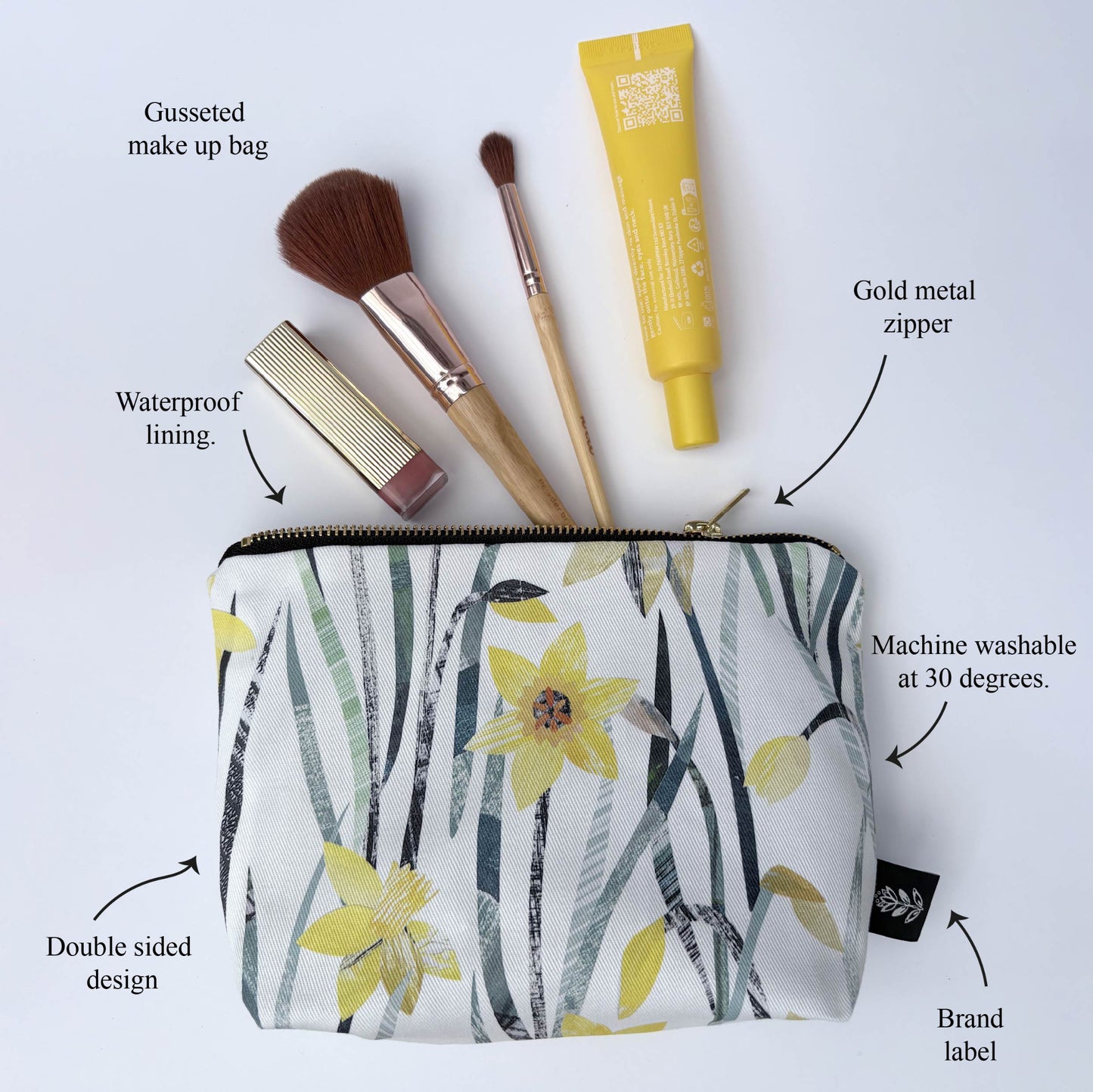 A Daffodil make up bag has been put on a white background and has two make up brushes and some lipstick and a tube of cream coming out of it.