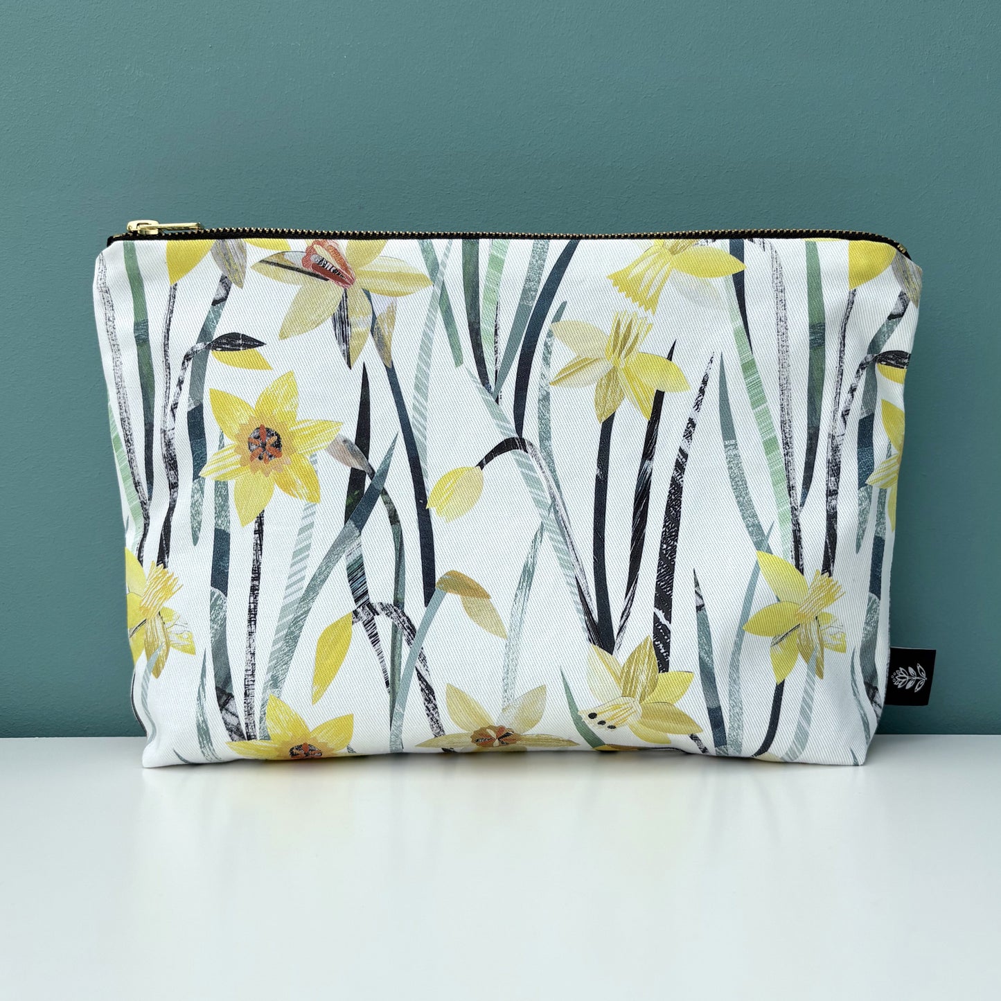 A Daffodil wash bag has been placed on a white shelf against a blue green backdrop.