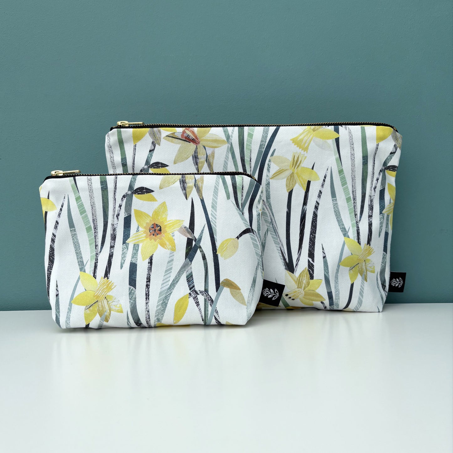 A daffodil make up bag has been placed in front of a daffodil wash bag.  They have been placed on a white shelf against a blue green back drop.