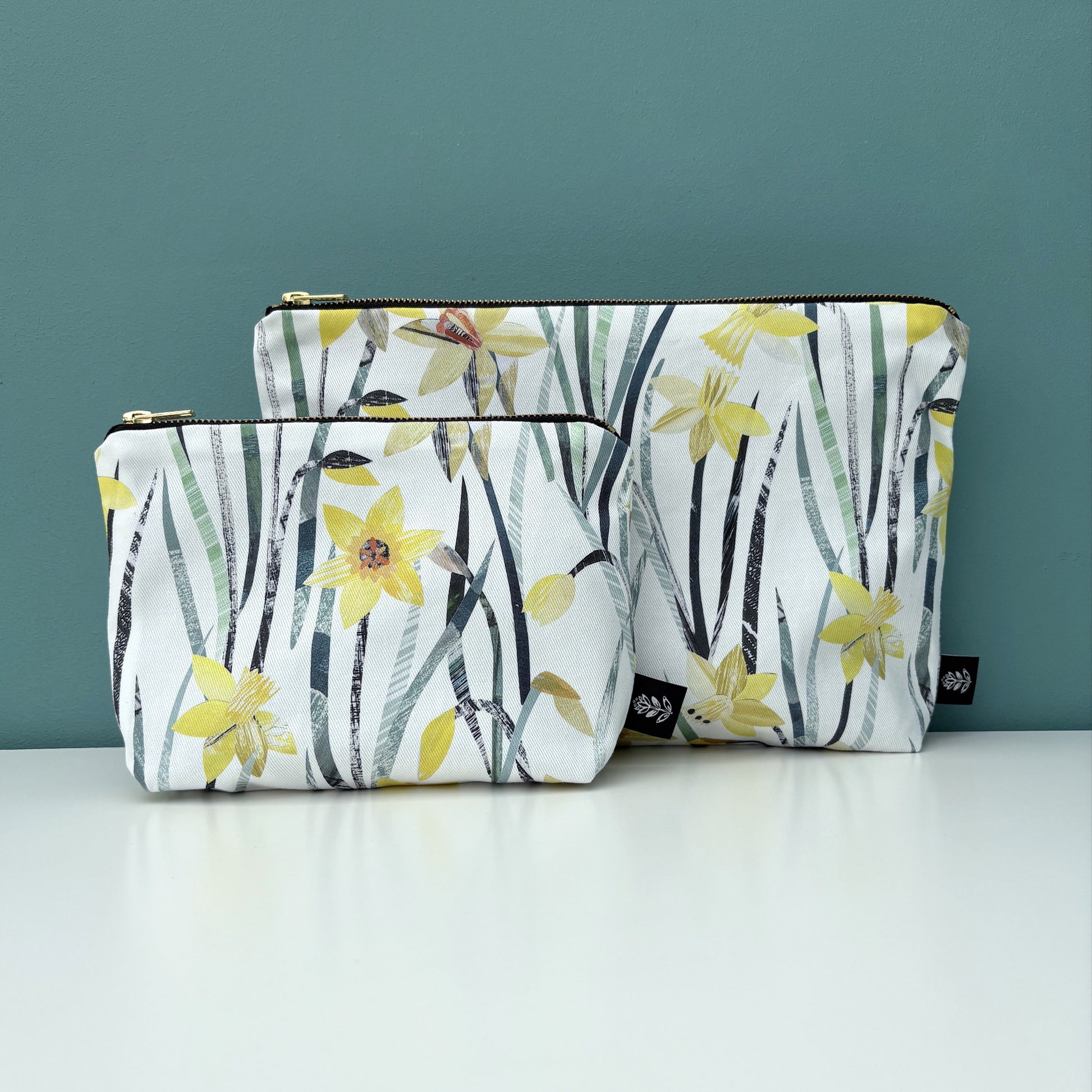 A daffodil make up bag has been placed in front of a daffodil wash bag.  They have been placed on a white shelf against a blue green back drop.