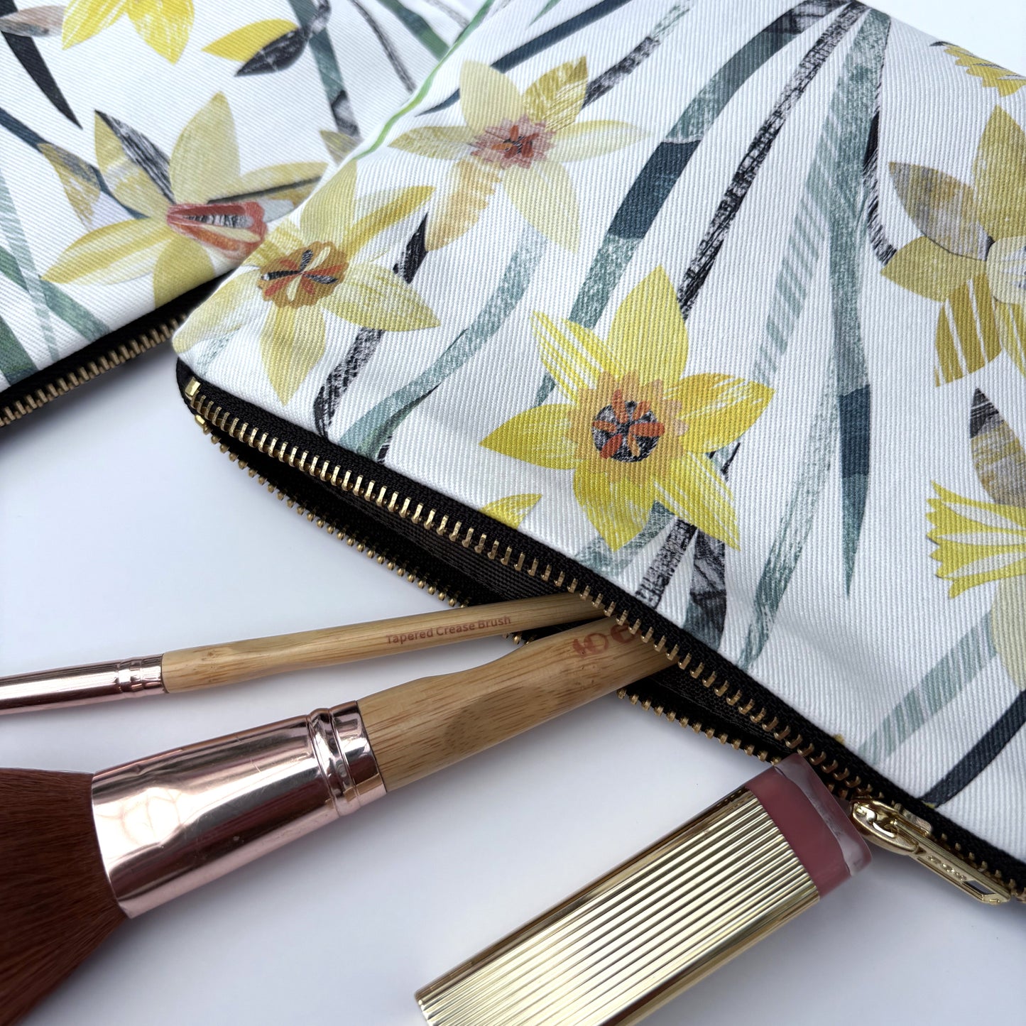 Close up of a daffodil wash bag featuring the pretty yellow and green daffodil flowers, you can see the gold zip and green lining. Two make up brushes and a lipstick are coming out of the wash bag.