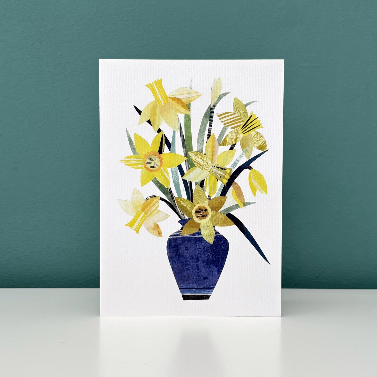 A greeting card featuring a blue vase of yellow daffodils has been stood on a white shelf against a Teal wall.