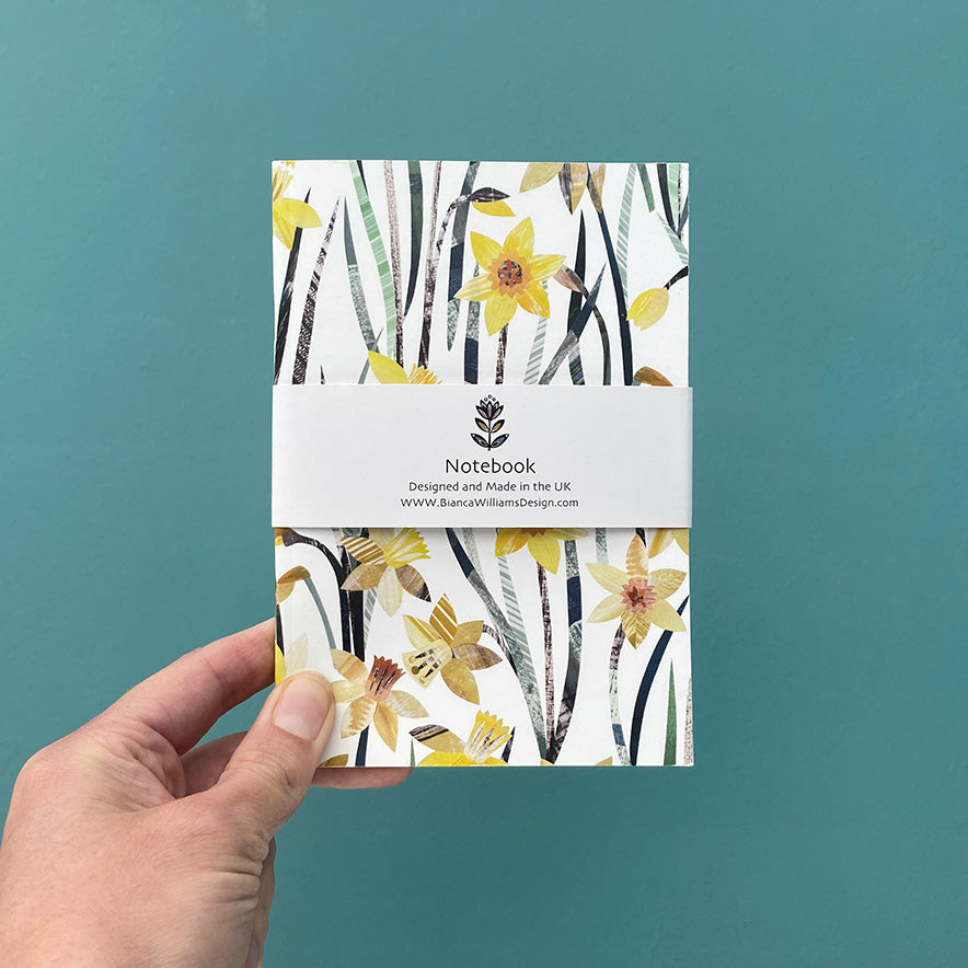 Small white notebook with all over Daffodil pattern packaged in a white branded belly band and held in front of a blue green back ground.