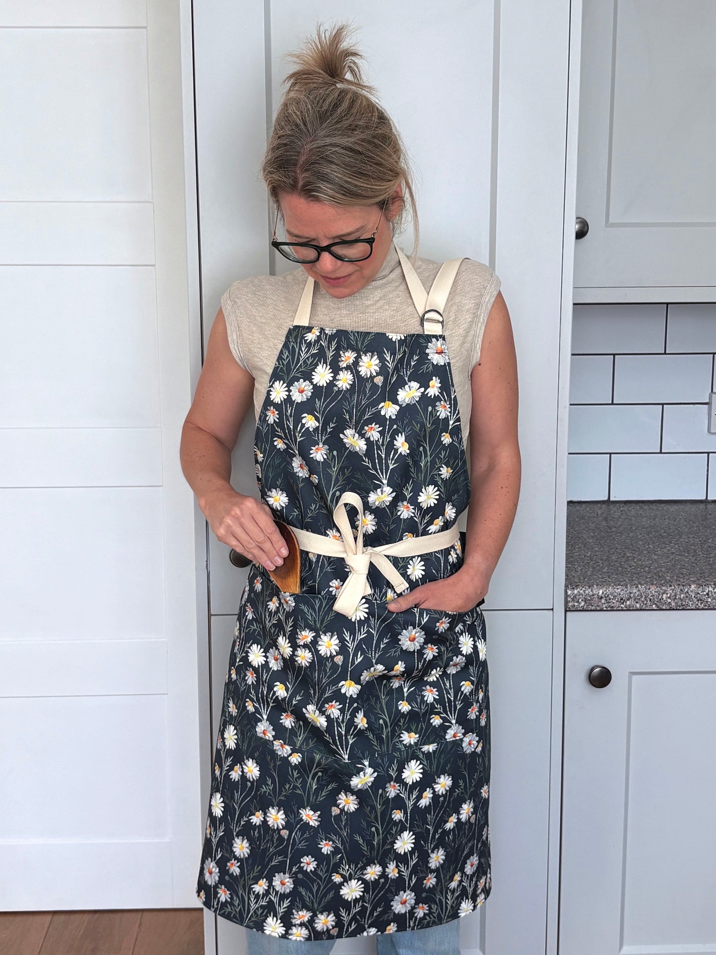 A lady is wearing the Daisy apron with two pockets on the front and an adjustable neck strap in a grey kitchen.  She has tied the apron around her waist.