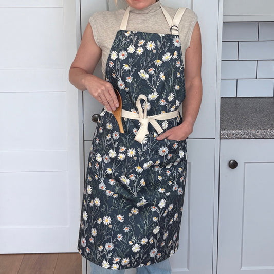 Woman wearing a white Daisy Apron on a Navy Blue background is standing in a pale grey kitchen.
