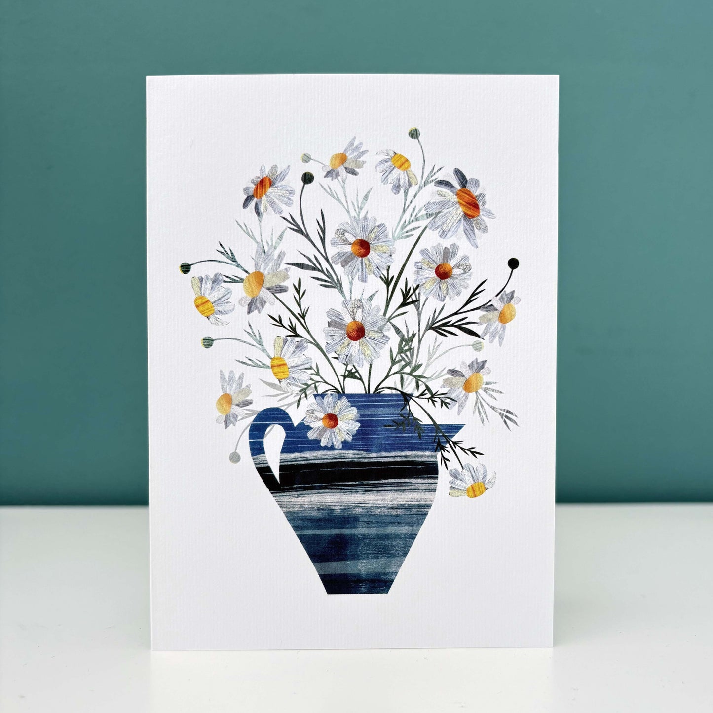 A white card featuring Daisies in a textured blue Vase.  It has been stood on a white surface with a Teal background.