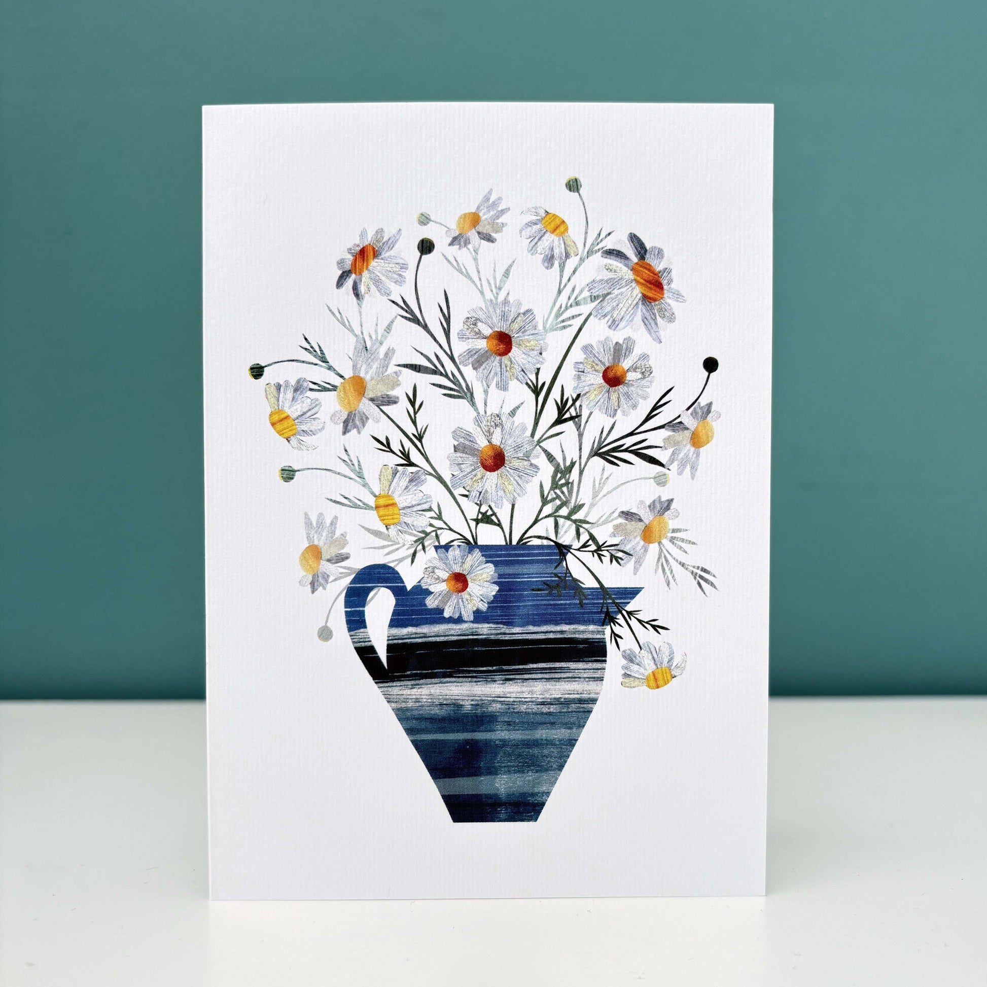 A white card featuring Daisies in a textured blue Vase.  It has been stood on a white surface with a Teal background.
