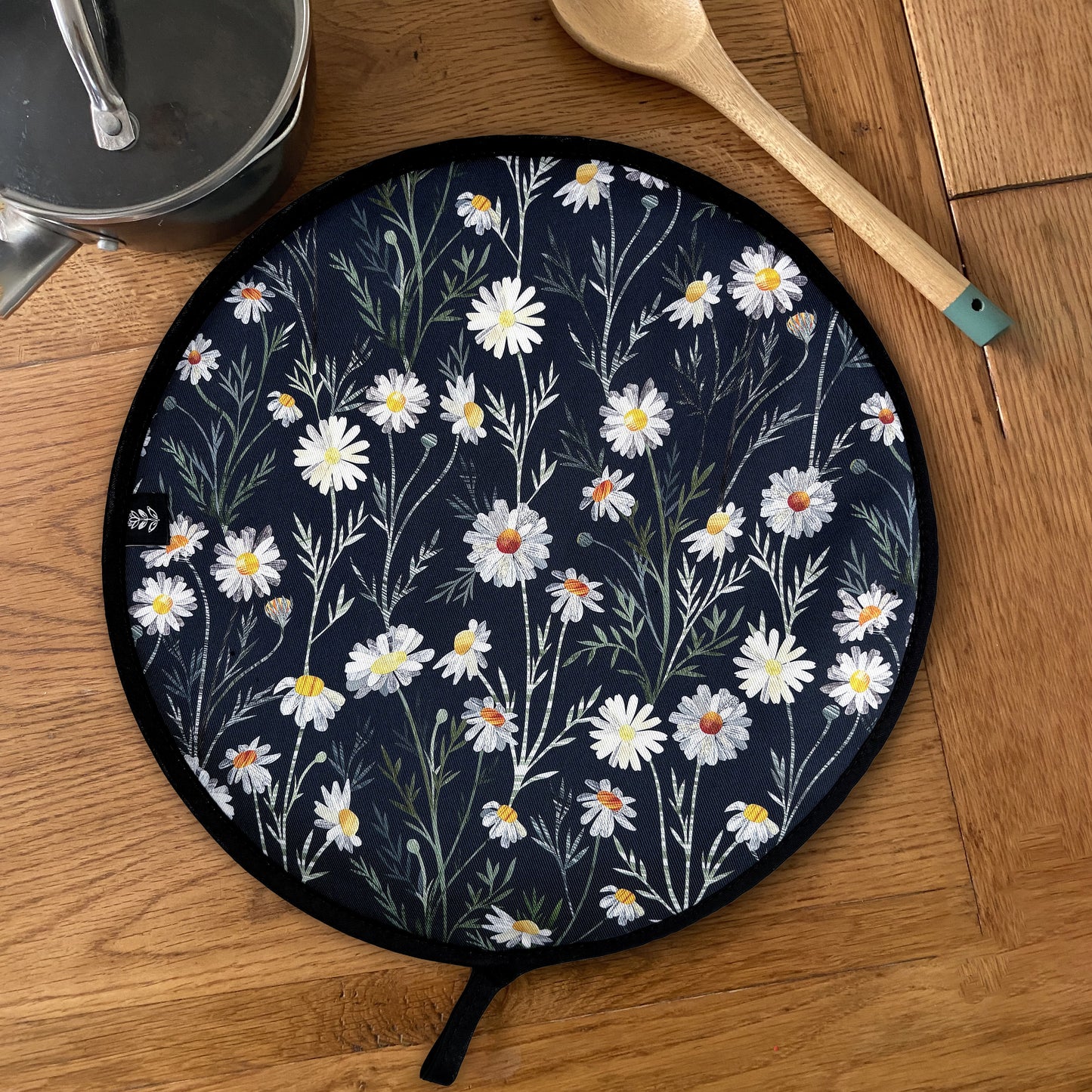 A Daisy Aga Cover has been placed on a wooden table with a saucepan and a wooden spoon. The Daisy pattern features pretty white daisy flowers on a dark Navy background.