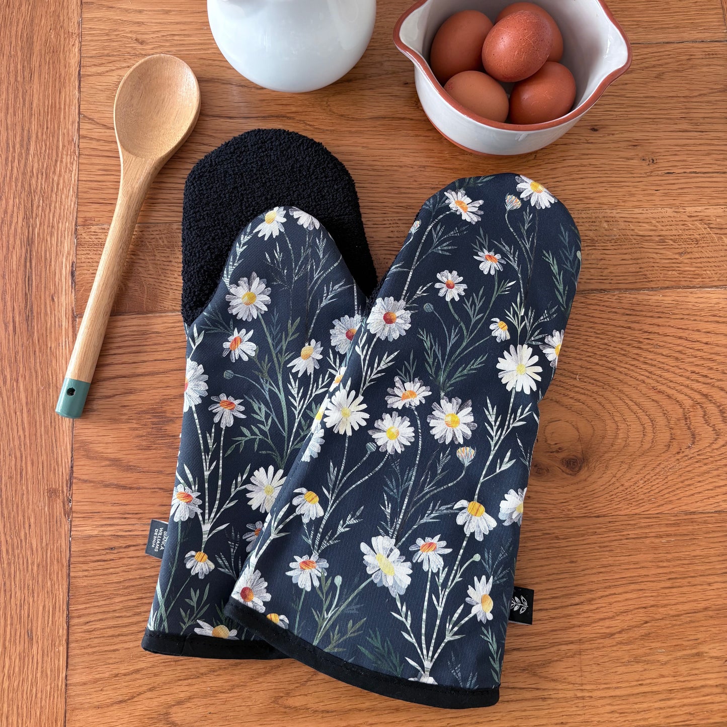 Two Daisy Floral-patterned oven mitts on a wooden surface with a wooden spoon, white pitcher, and bowl of eggs.