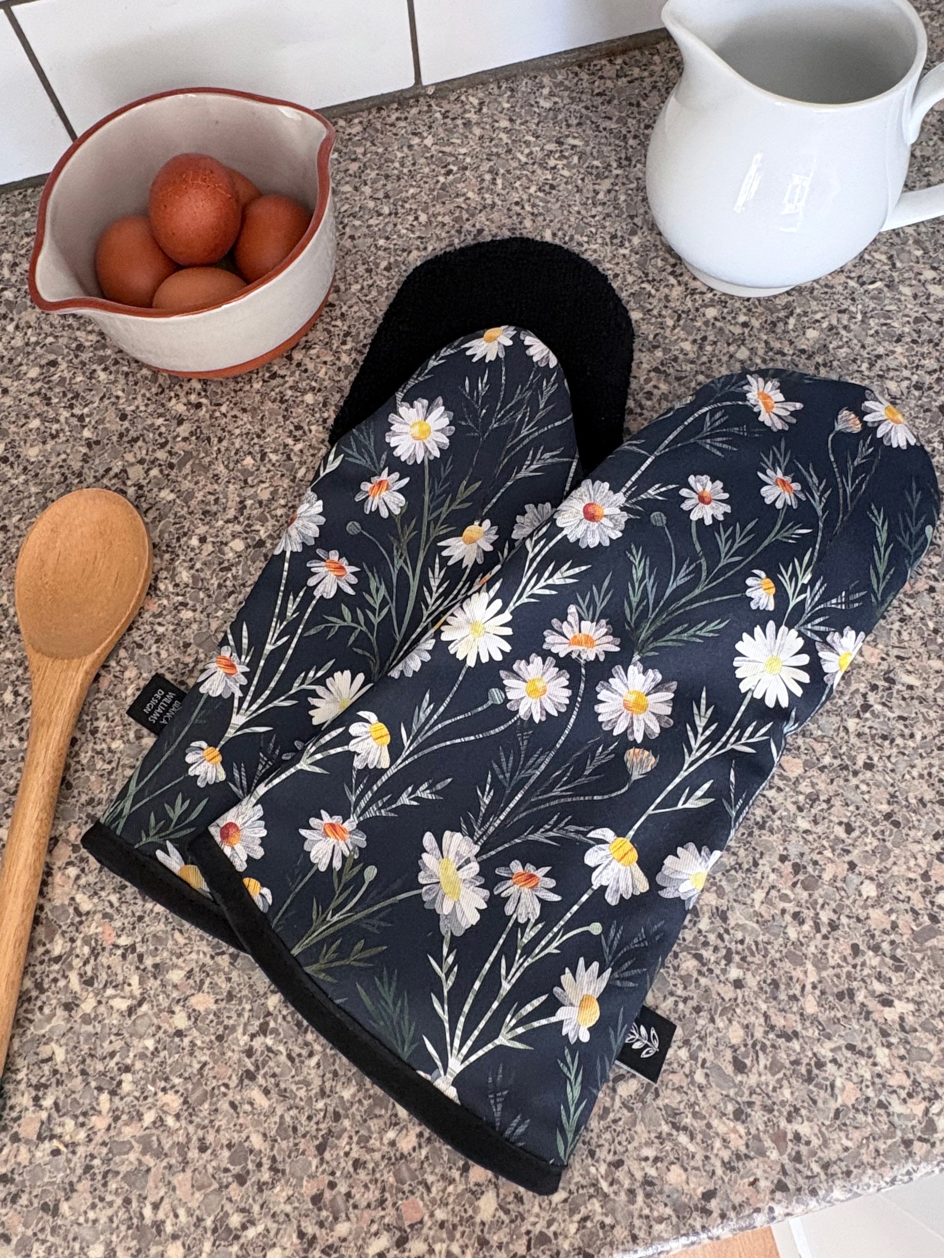 Navy Daisy Floral-patterned oven mitts on a kitchen counter with a wooden spoon and ceramic bowl.