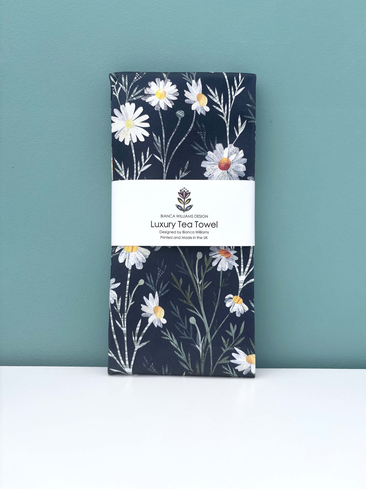A Daisy Tea Towel has been packaged in a branded belly band and placed on a white shelf against blue green wall.