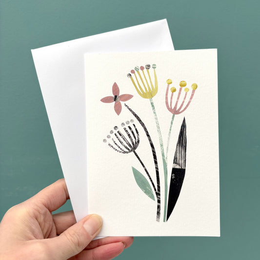 A hand holds a white greeting card featuring a minimalist botanical illustration of flowers and leaves in muted pink, yellow, green, and black, with a white envelope behind it against a solid teal background.
