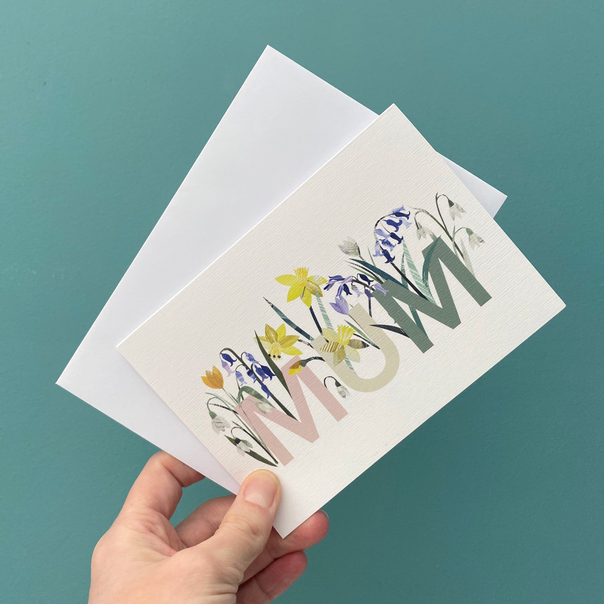 Floral Mum card is being held with a white envelope against a blue green background.