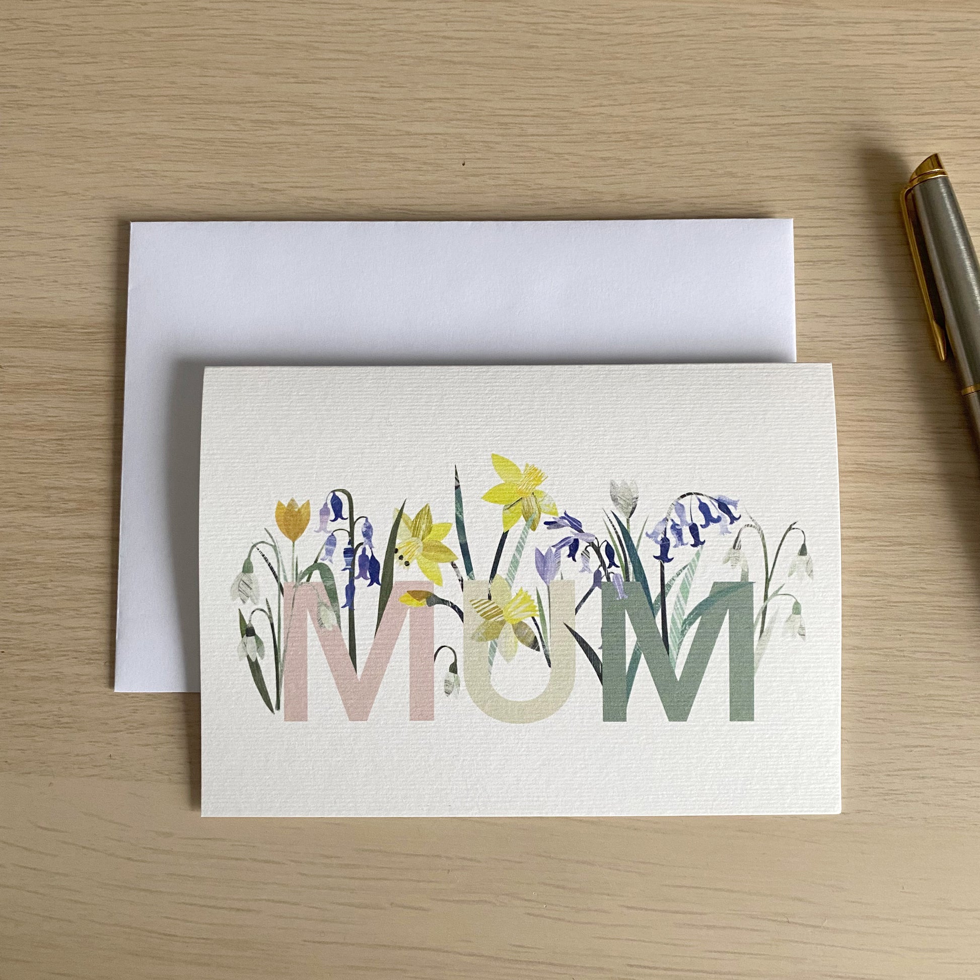 Mum card has been placed flat on a wooden table with a white envelope and a pen beside it.