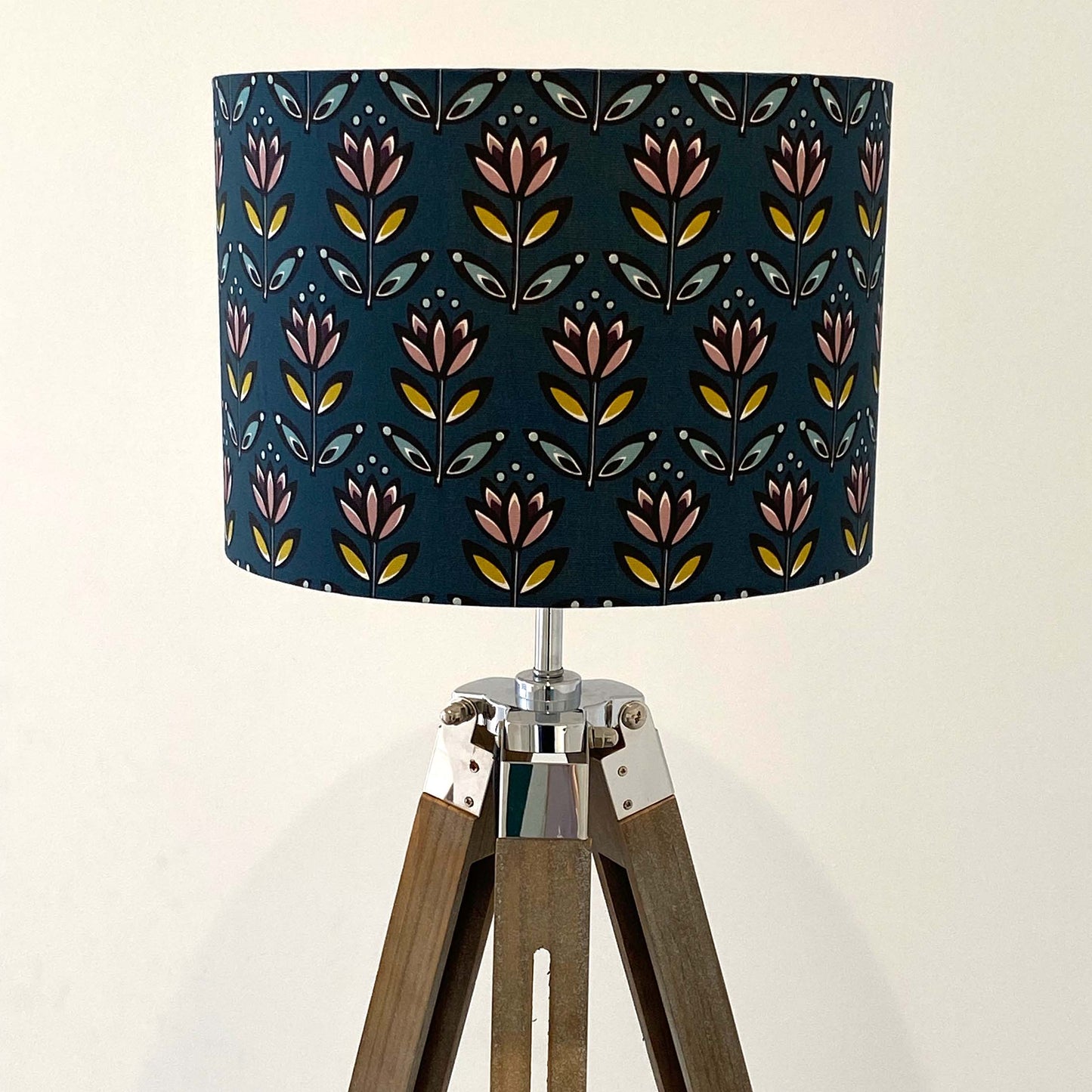 Medium Teal Flower Lampshade displayed here on a wooden tripod floor lamp. The design features a simplified flower motif in black, white, yellow, blue, dusky pink and aubergine on a Teal background. The light is shown off in this photograph.