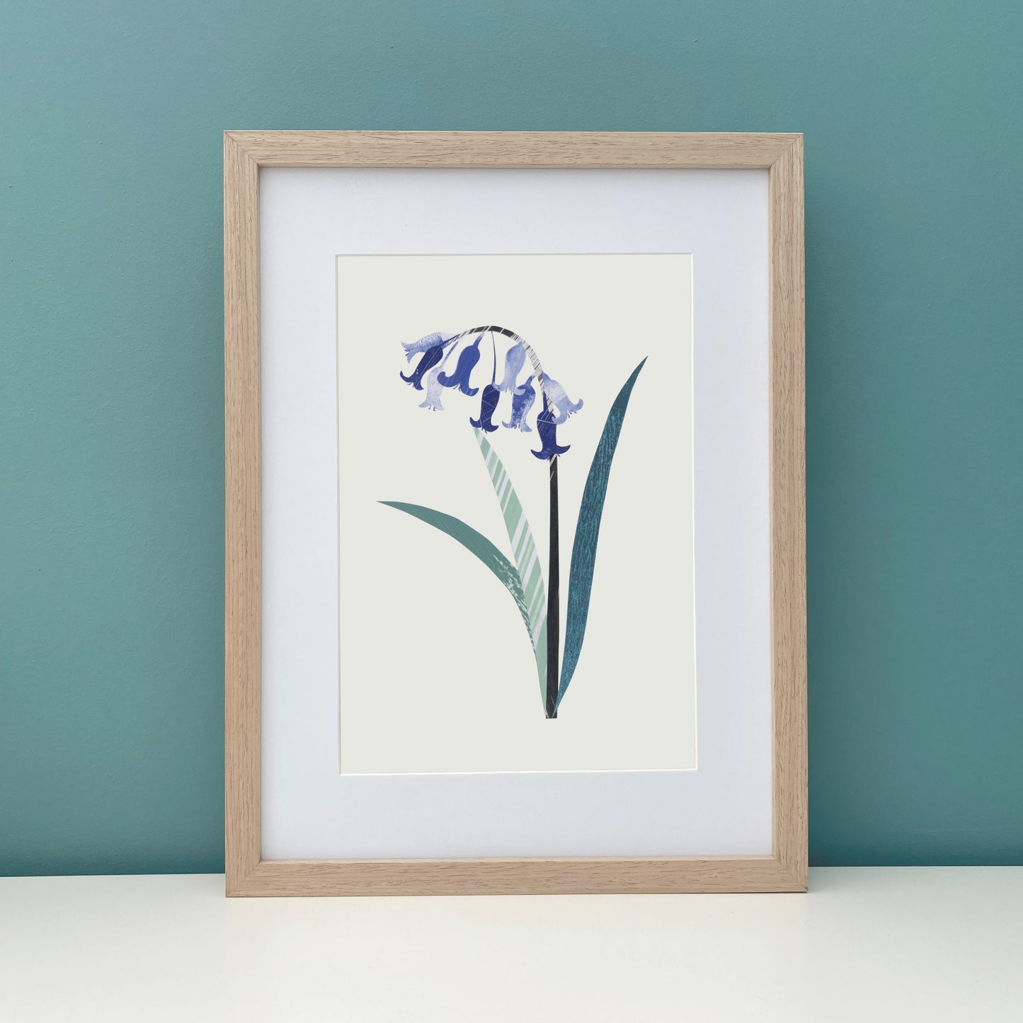 Framed artwork of a bluebell flower on a off white background has been hung on a teal wall.