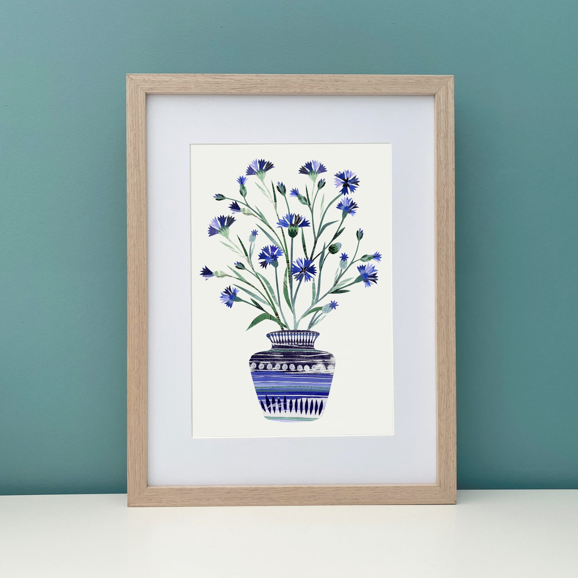 Framed artwork of a blue Cornflowers floral design on a white background against a teal wall.