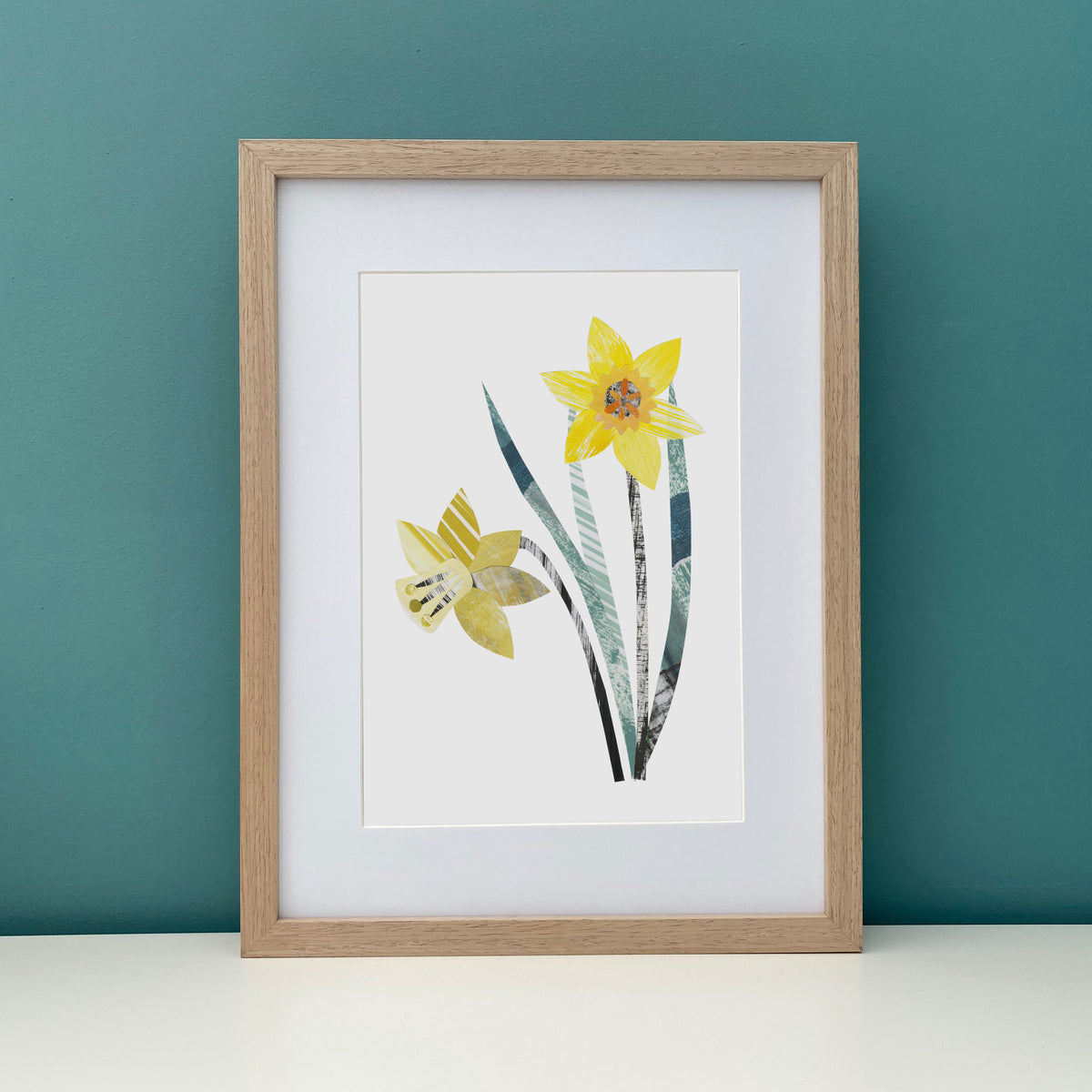 Framed artwork of a A4 print of a Yellow Daffodil Flower.  It has been put on a white shelf against a Teal Wall.
