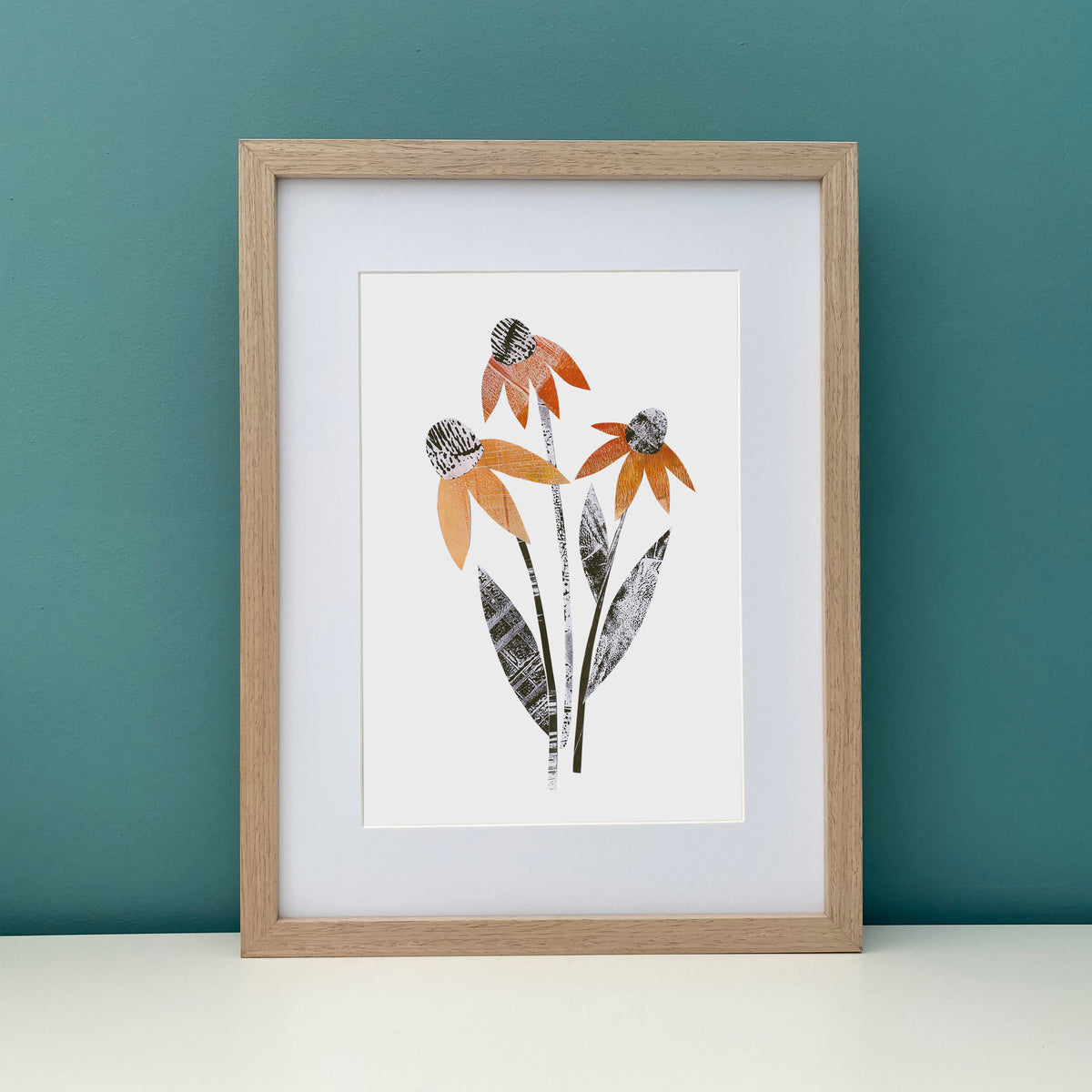wooden framed artwork of a stylised orange rudbeckia flower.  The framed artwork has been placed on a white shelf against a tea all.