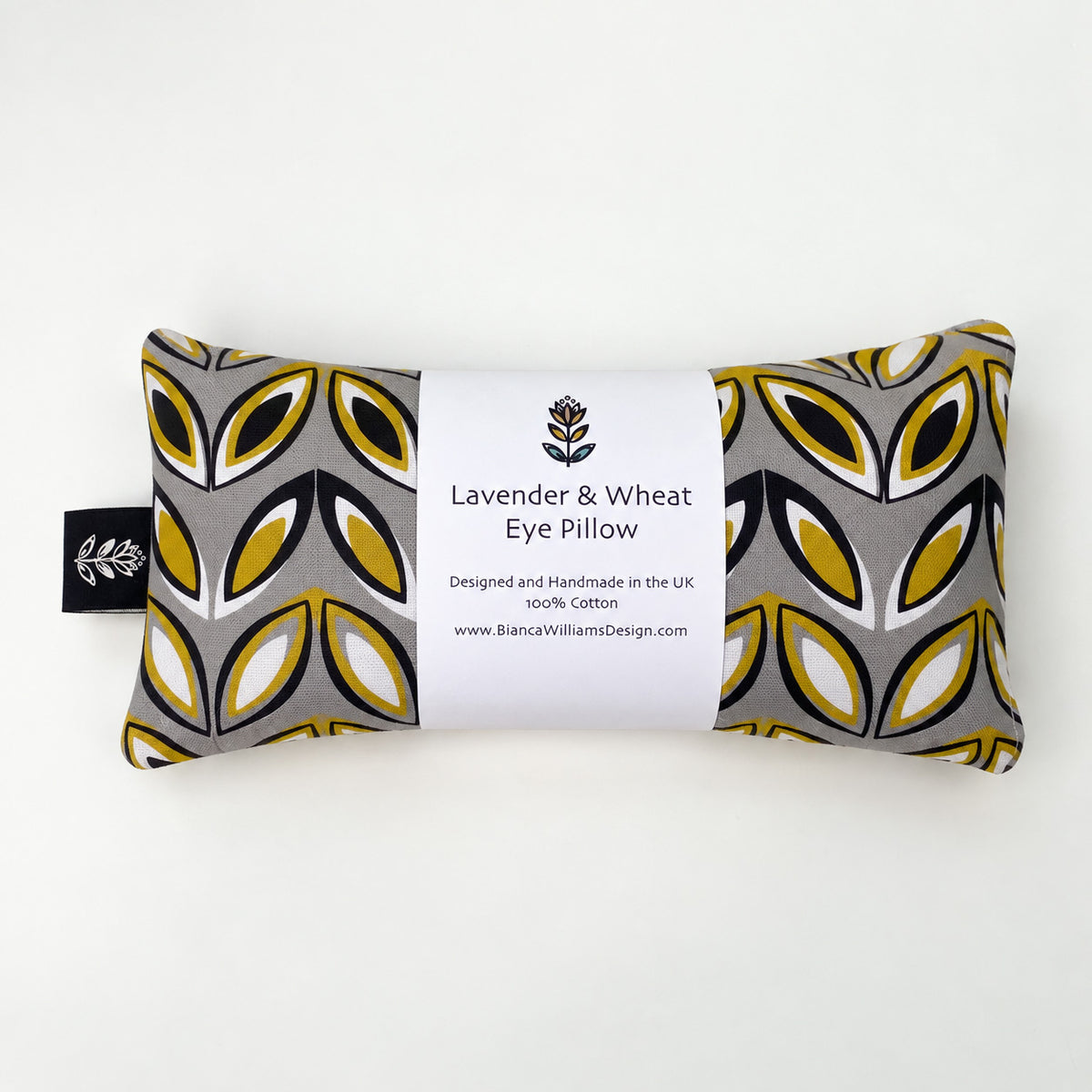 A rectangular lavender and wheat eye pillow with a grey, yellow, and black geometric pattern, wrapped in a white paper label that reads 'Lavender & Wheat Eye Pillow' and 'Bianca Williams Design'.
