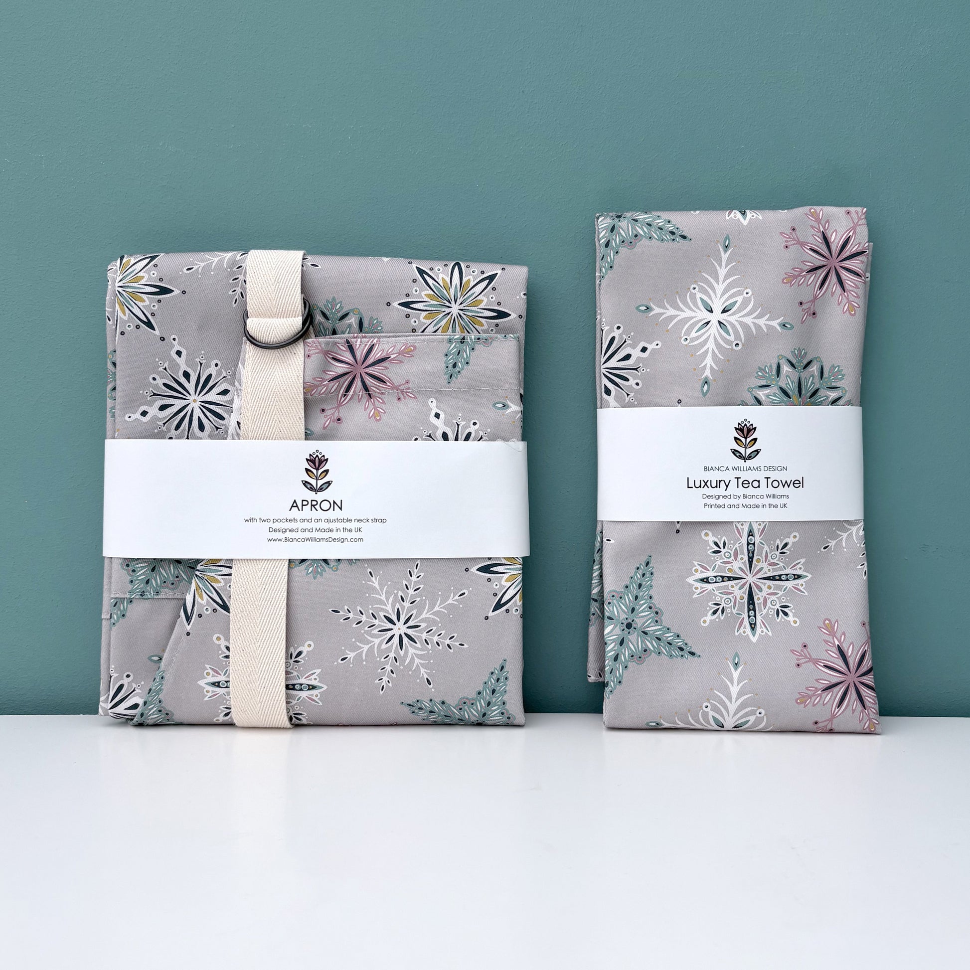 A matching grey snowflake apron and tea towel have been folded and packaged in a branded belly band and they have been placed on a white shelf against a blue green background.