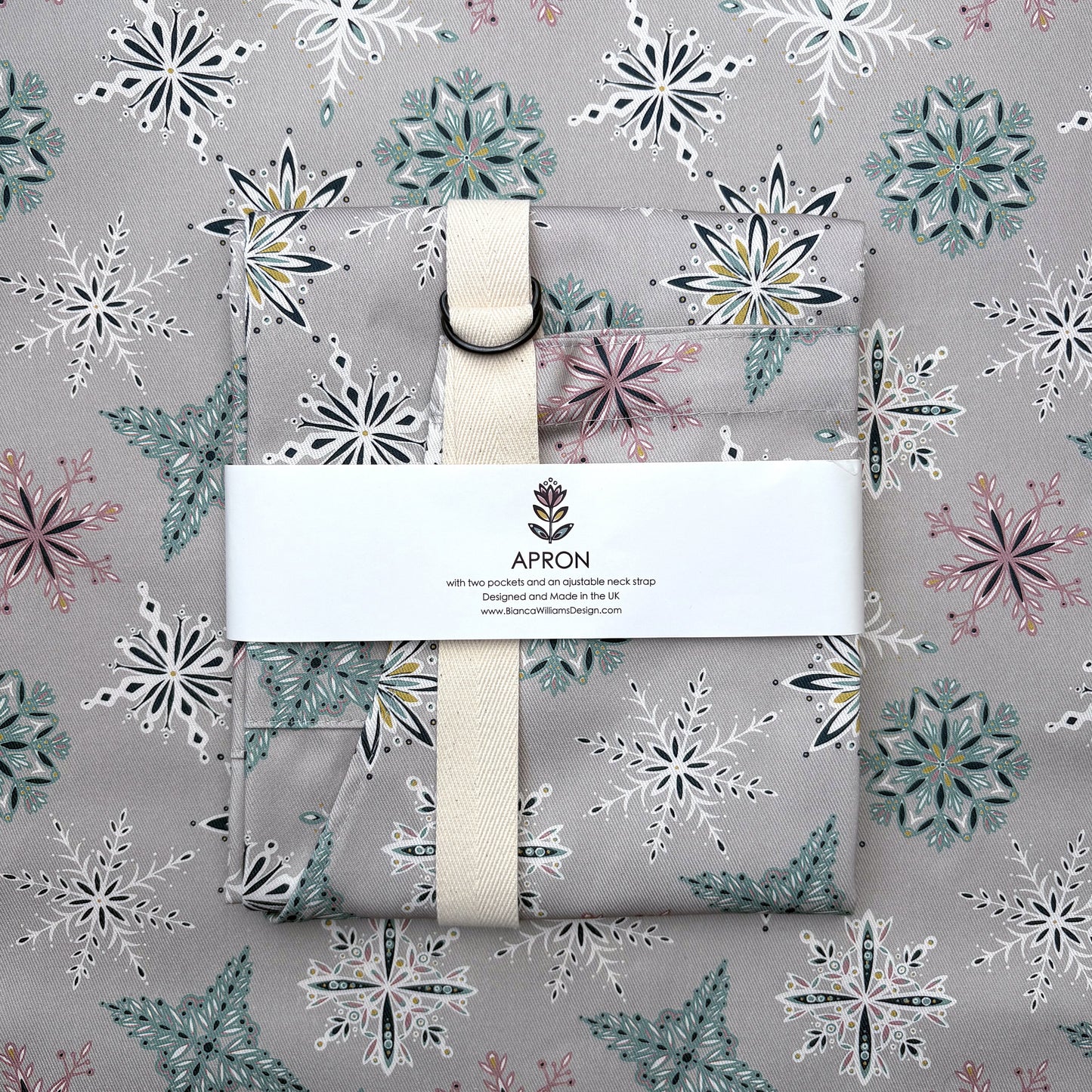 A grey snowflakes apron has been folded and wrapped in a branded belly band and has been placed on a piece of grey snowflakes patterned fabric.