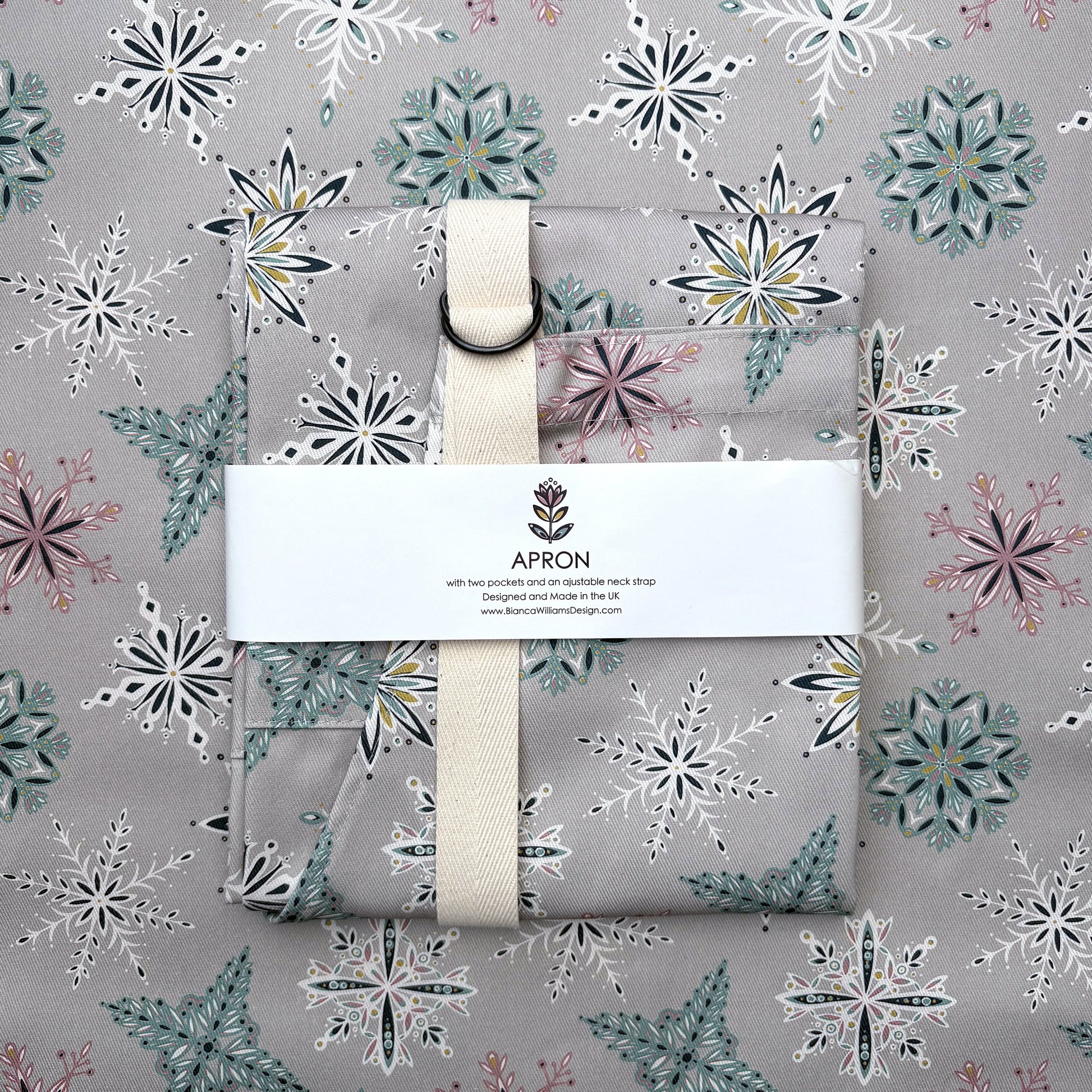 A grey snowflakes apron has been folded and wrapped in a branded belly band and has been placed on a piece of grey snowflakes patterned fabric.