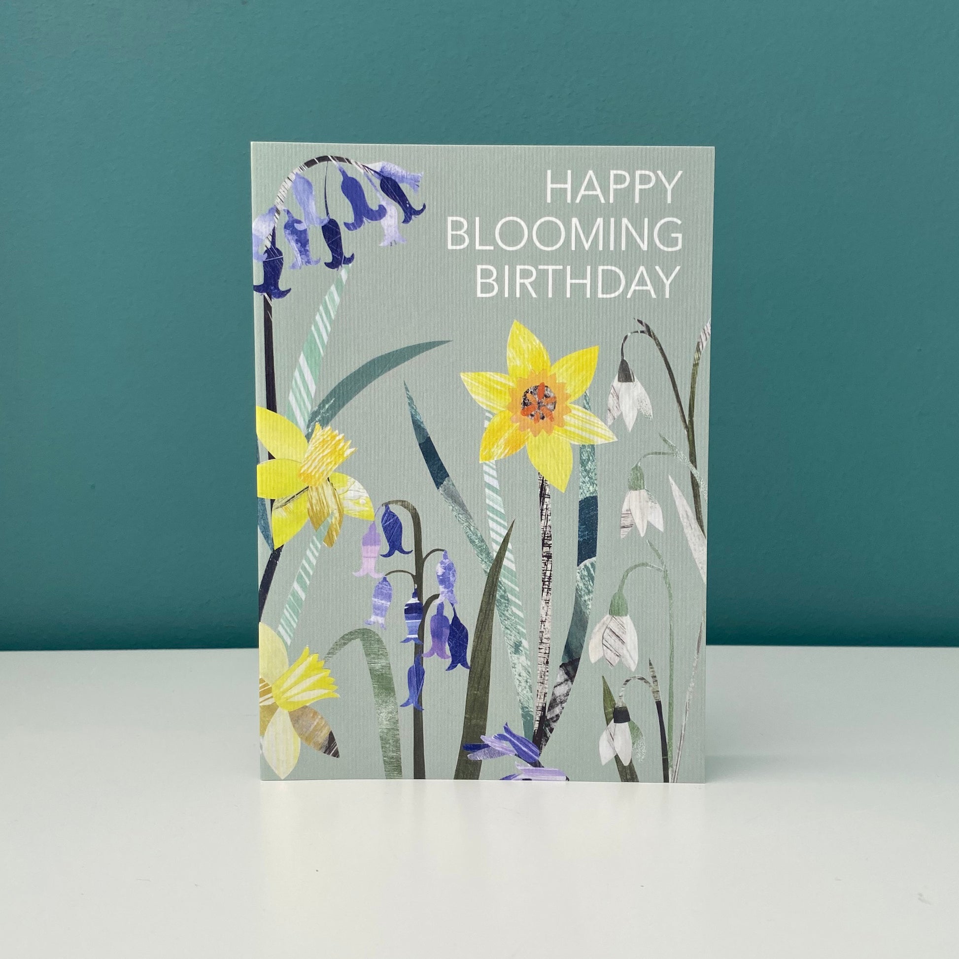 A birthday card with the words Happy Blooming Birthday on it features collaged Daffodil, Bluebell and snowdrop flowers on a soft sage green background.  The card is stood on a white shelf against a blue green wall.