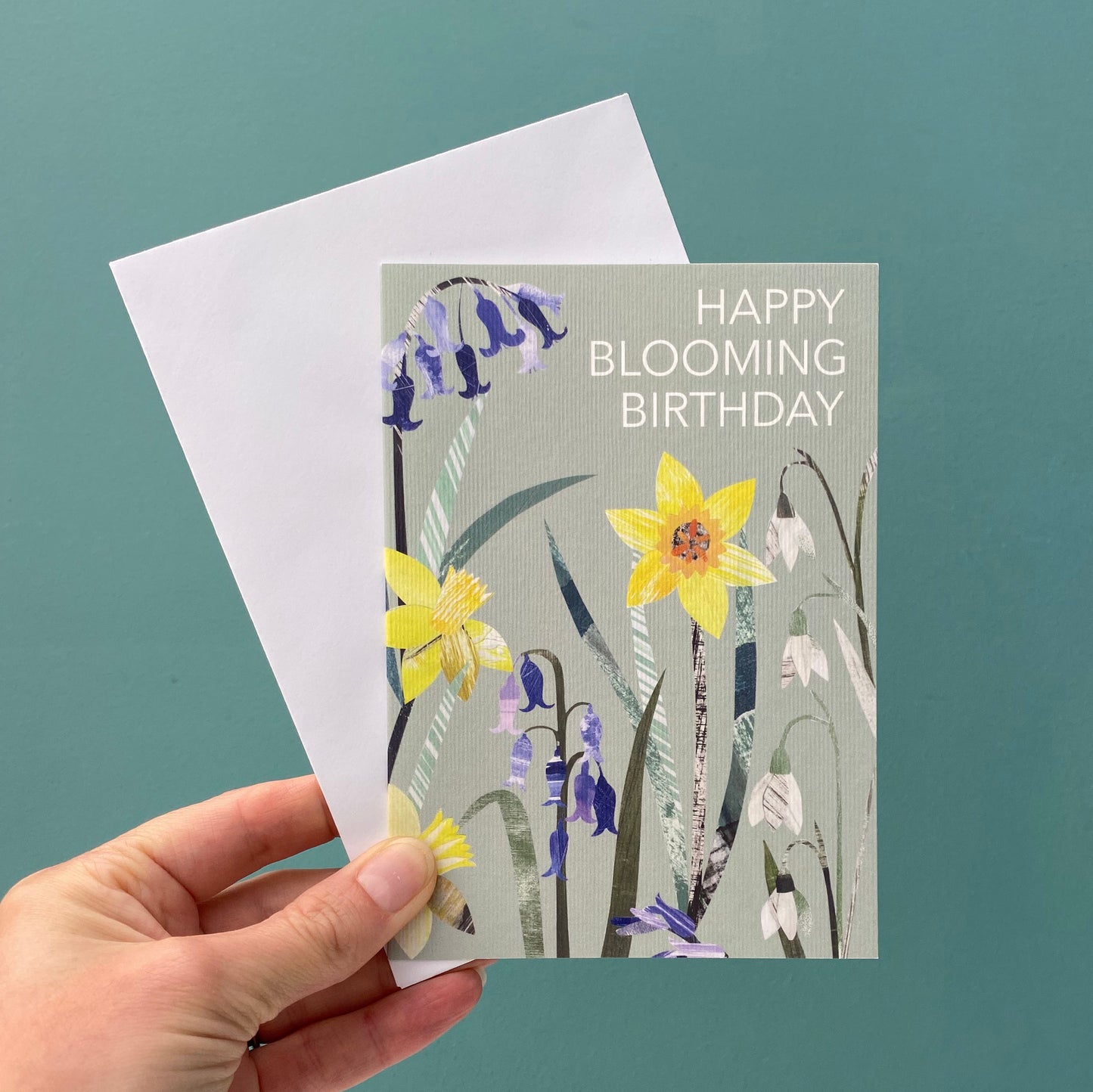 A birthday card with the words Happy Blooming Birthday on it features collaged Daffodil, Bluebell and snowdrop flowers on a soft sage green background.  The card is being held with a white envelope against a blue green background.