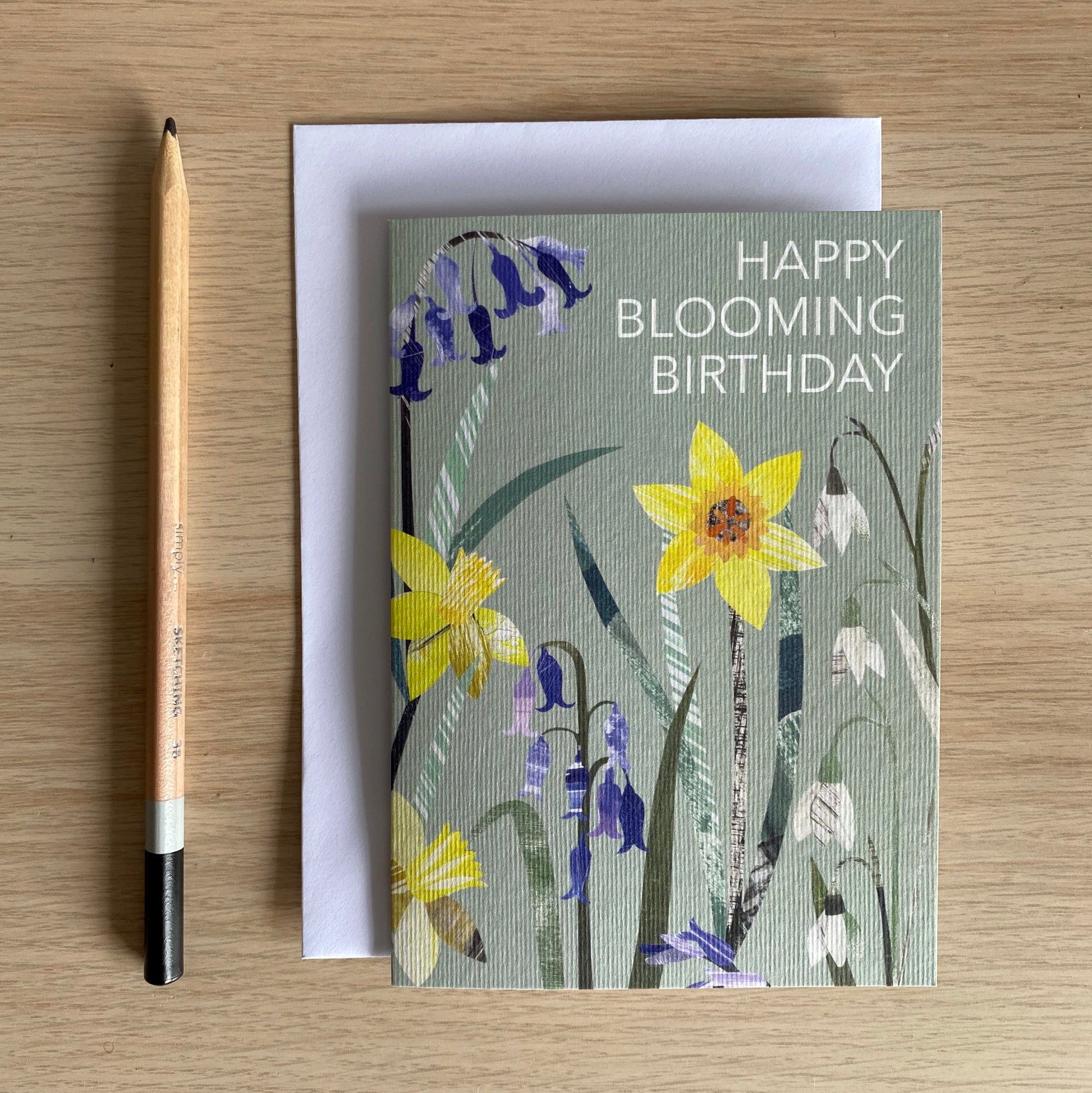 The Happy Blooming Birthday Card has been placed on a wooden table with a plain white envelope and pencil.