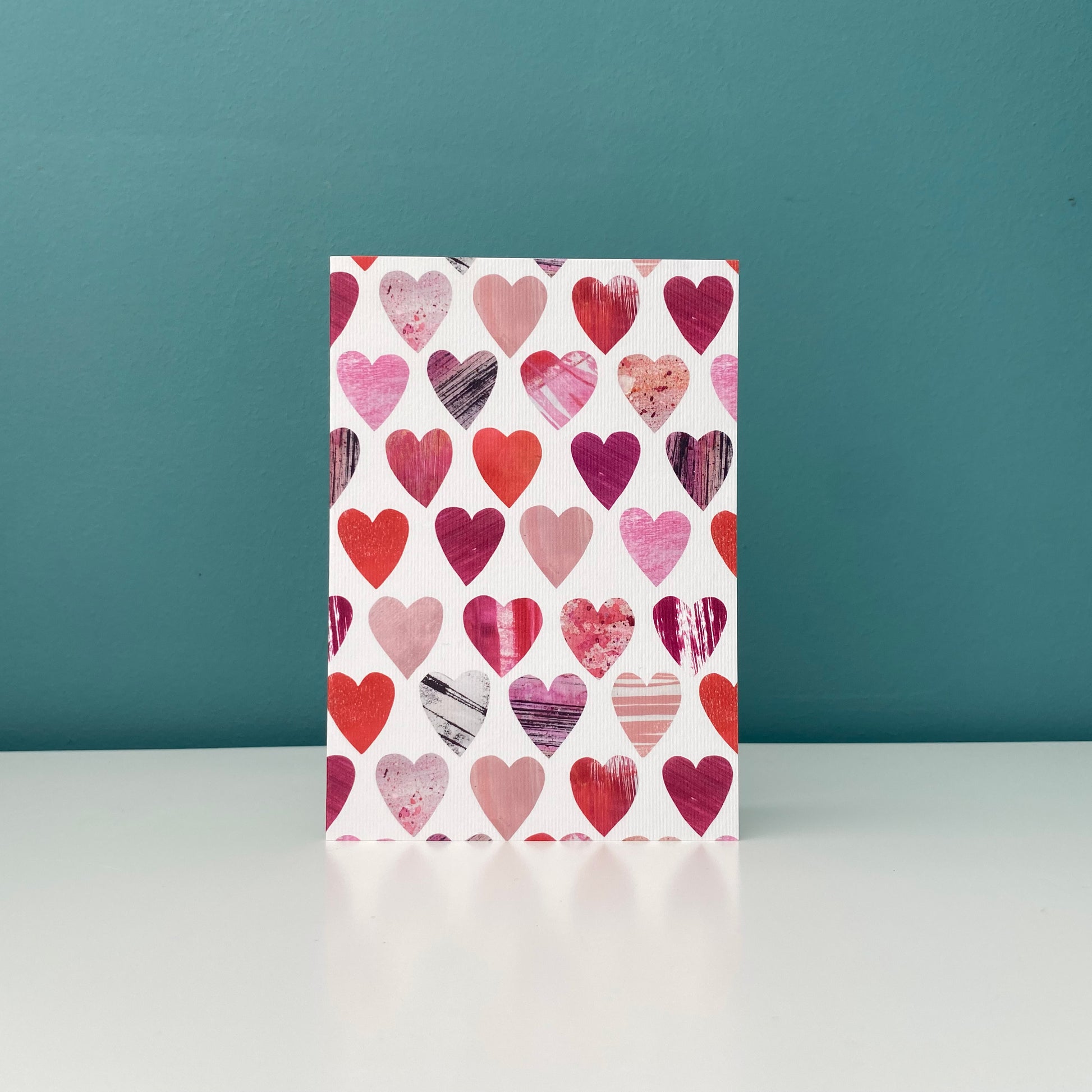 A card featuring all over textured Pink and Red Heart motifs on a white background has been stood up on a white shelf with a blue green wall behind it.