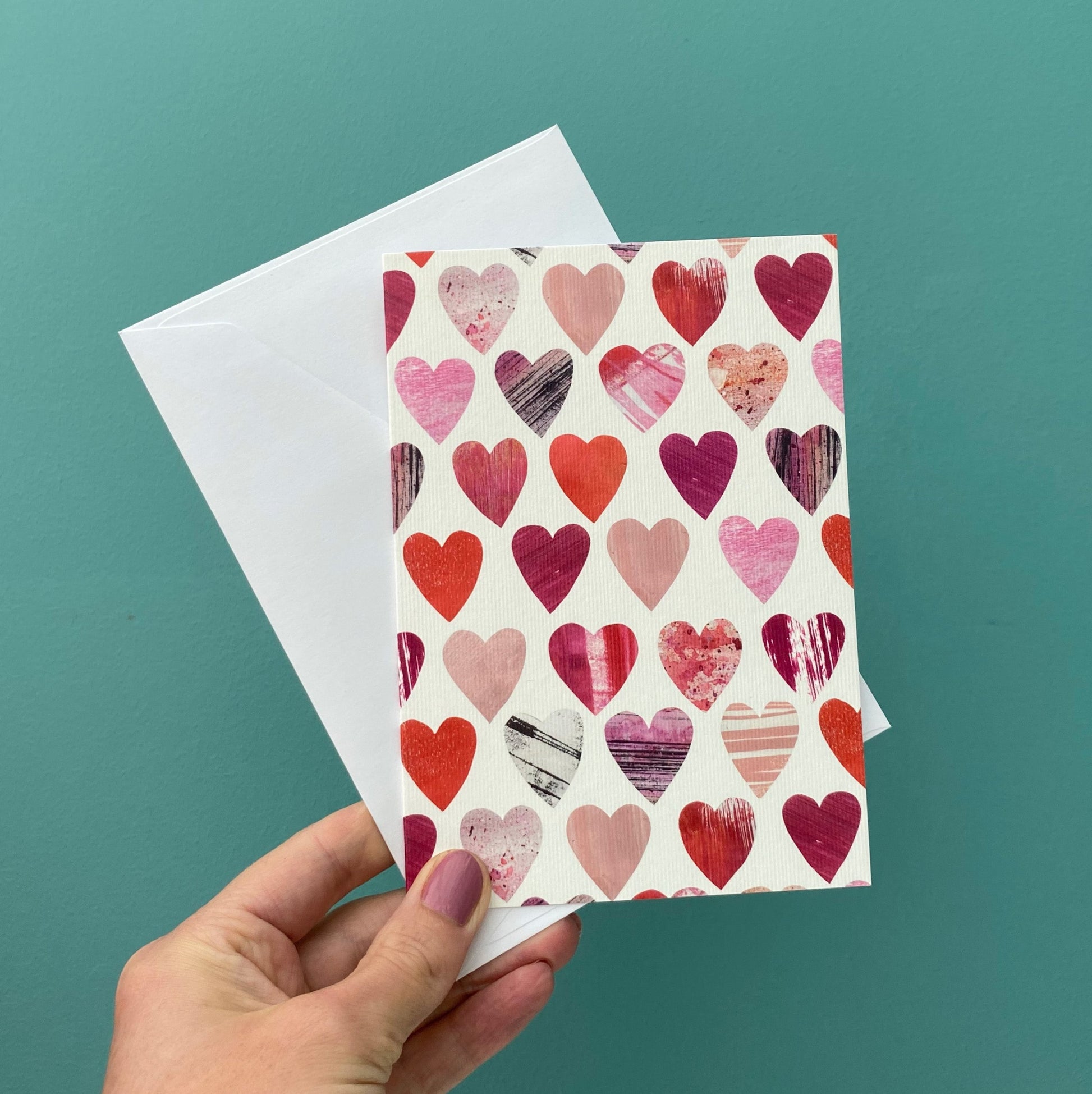 A card featuring all over textured Pink and Red Heart motifs on a white background, is being held with a white envelope against a blue green wall.