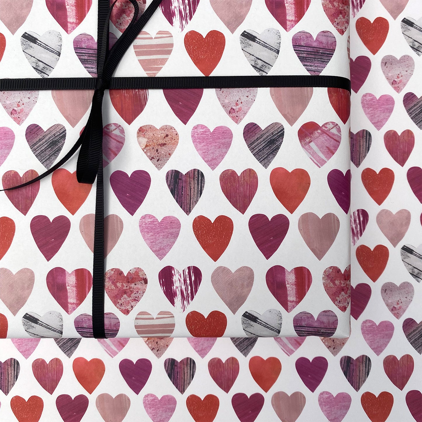 A close up of a present wrapped in the heats wrapping paper.  It has been tied with black ribbon and placed on a sheet of the same texture heart wrapping paper.