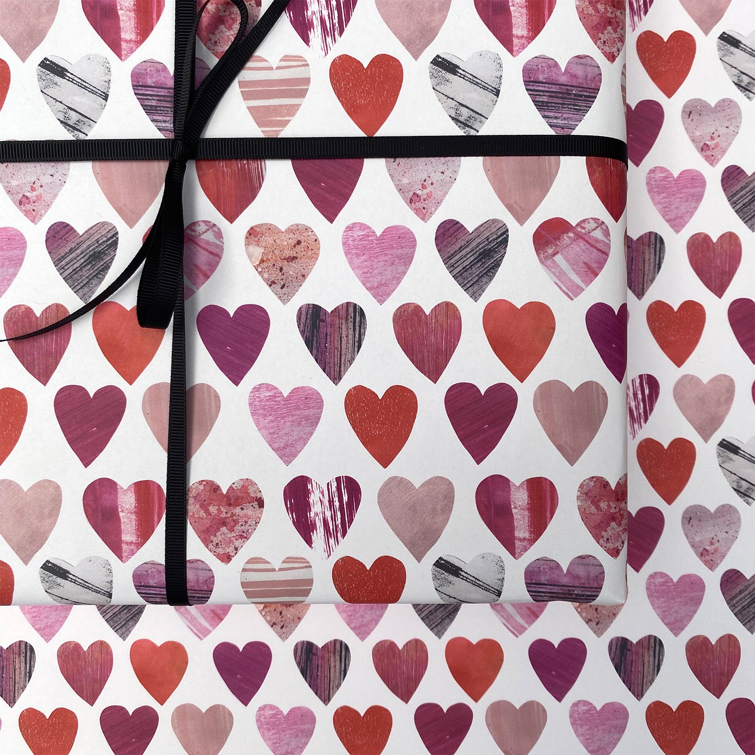 A close up of a present wrapped in the heats wrapping paper.  It has been tied with black ribbon and placed on a sheet of the same texture heart wrapping paper.