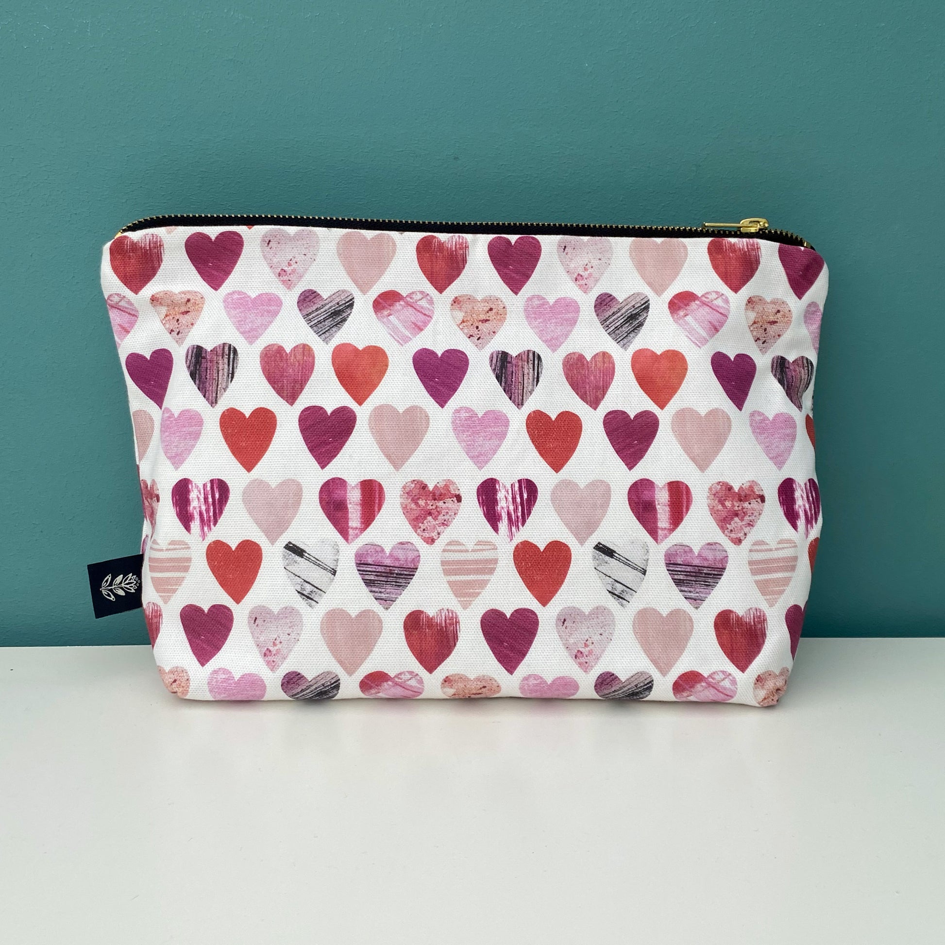 A large Hearts wash bag placed on a white shelf with a blue green background.  The wash bags has a gold metal zipper and a Brand label sewn into the outside seam.