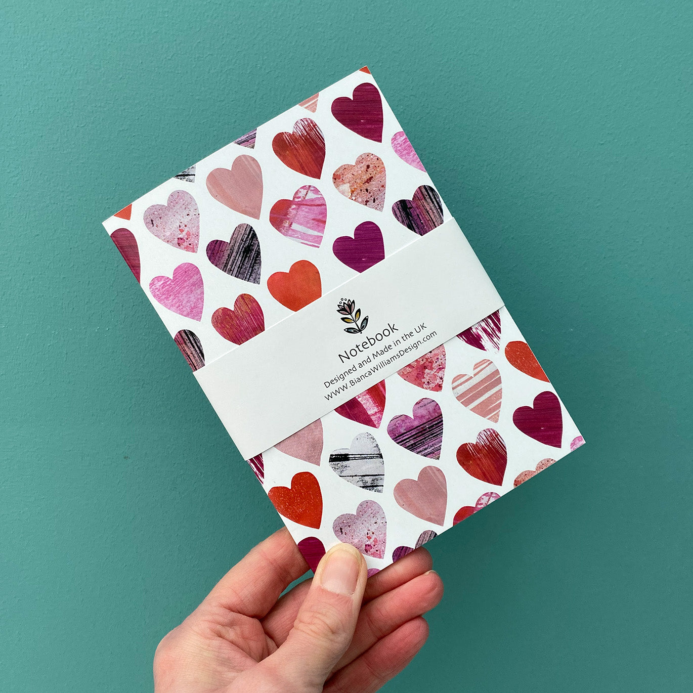 Small Hearts Notebook is being held up in front of a Blue Green wall.  The Notebook features a textured Pink and red heart pattern on a white ground.