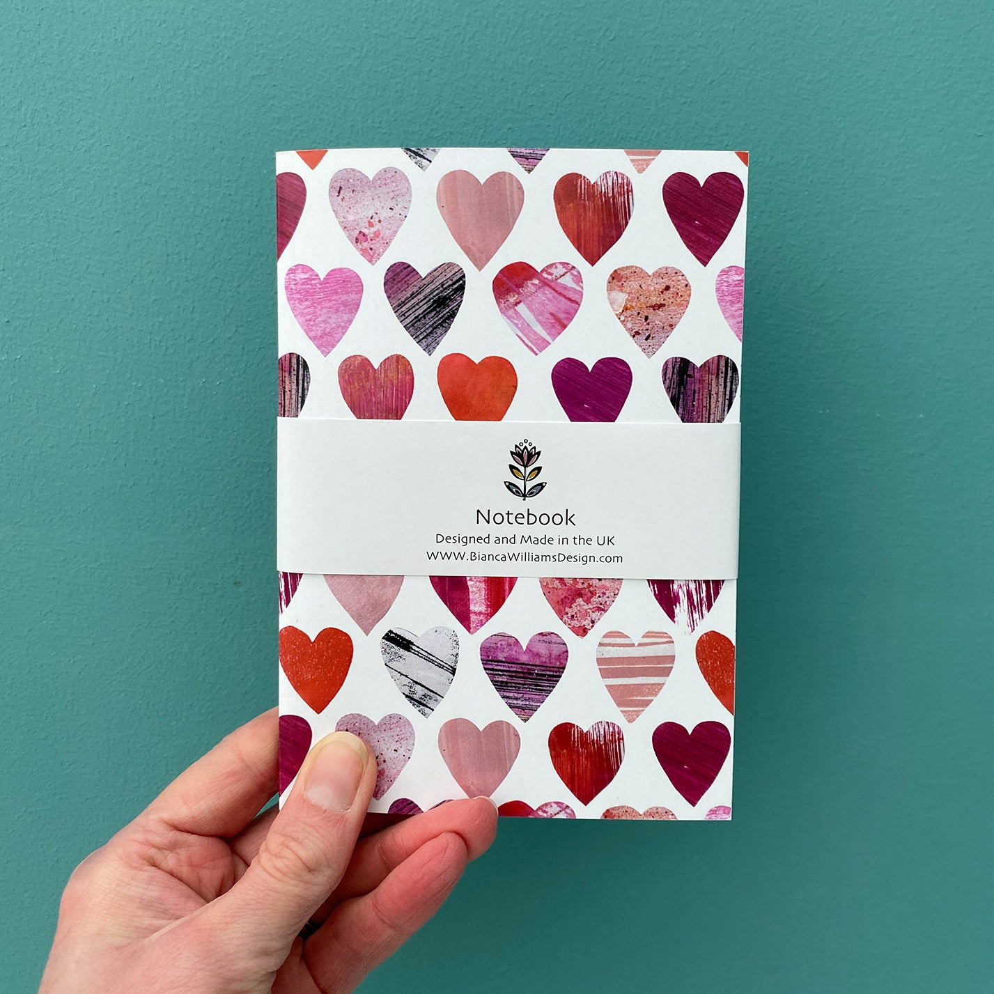 Small white notebook with all over pink and red heart pattern, packaged in white belly band and held in front of a blue green background.